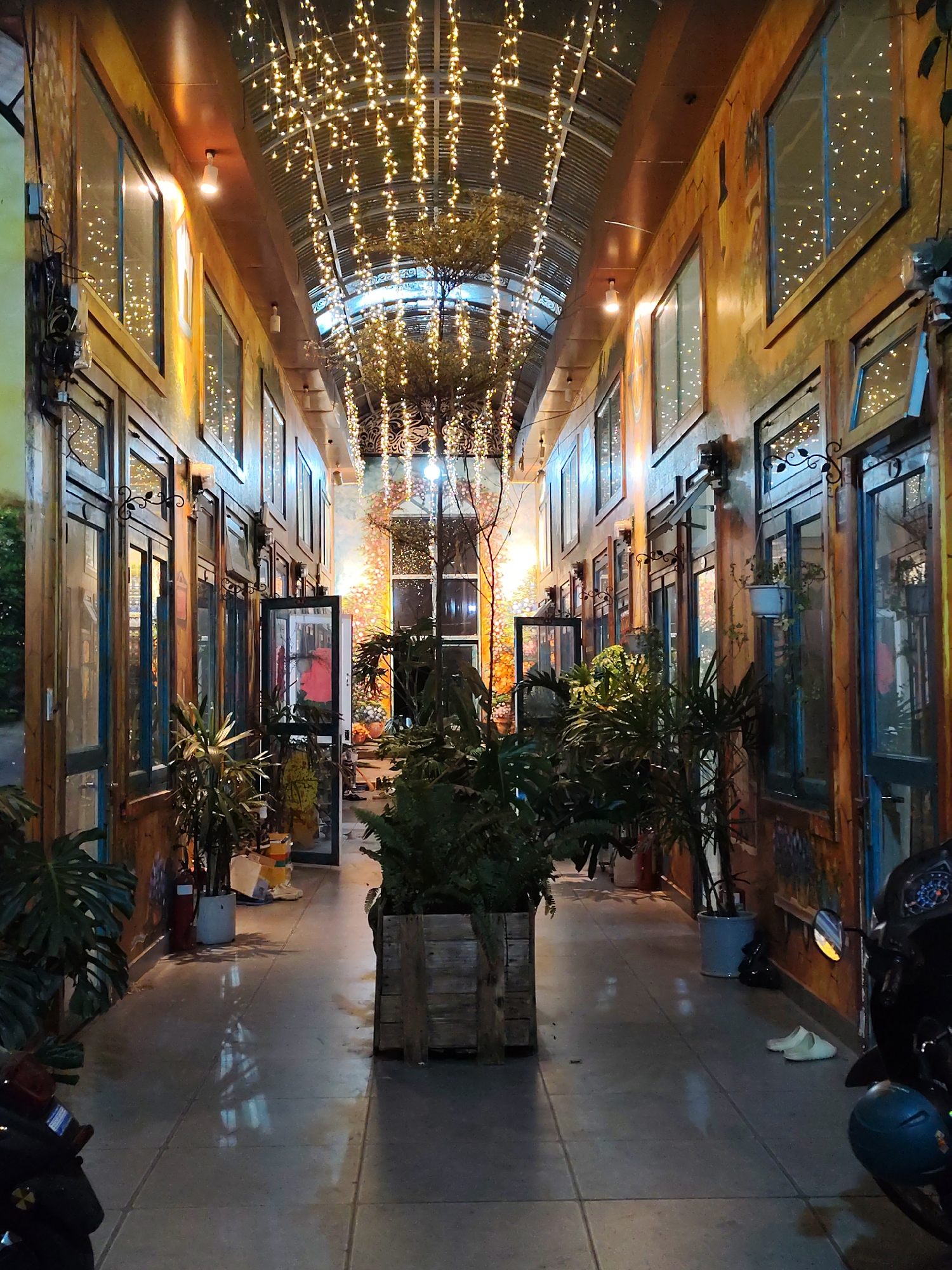 The image shows a long, covered walkway in Da Lat, Vietnam, decorated with strings of fairy lights hanging from the ceiling. The walkway is flanked by buildings on either side with multiple glass-paneled doors and windows. The walls of the buildings are decorated with murals or painted in a rustic style. Several potted plants are placed along the walkway. At the end of the walkway is an open area with a mural. A parked scooter is visible on the right side of the image. The floor of the walkway is tiled. The overall lighting creates a warm and inviting atmosphere.