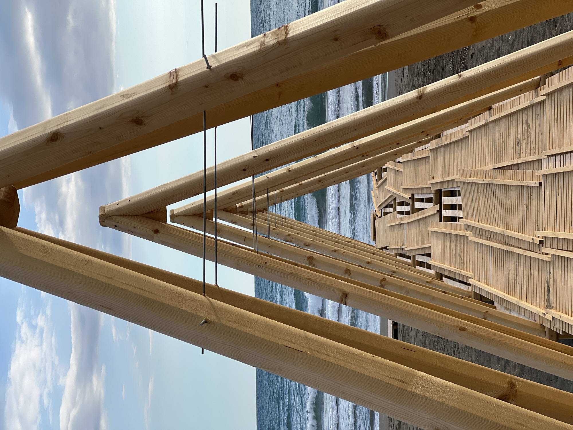 The image shows a partially constructed wooden structure near the sea. It appears to be a boardwalk or viewing platform, with wooden planks forming the base and wooden beams creating an overhead framework. Metal rods hang vertically from the beams, possibly for support or aesthetic purposes. The sea is visible in the background, with waves breaking on the shore. The sky is partly cloudy. This structure can be found at the beach in Ulcinj - Ulqin, Montenegro.