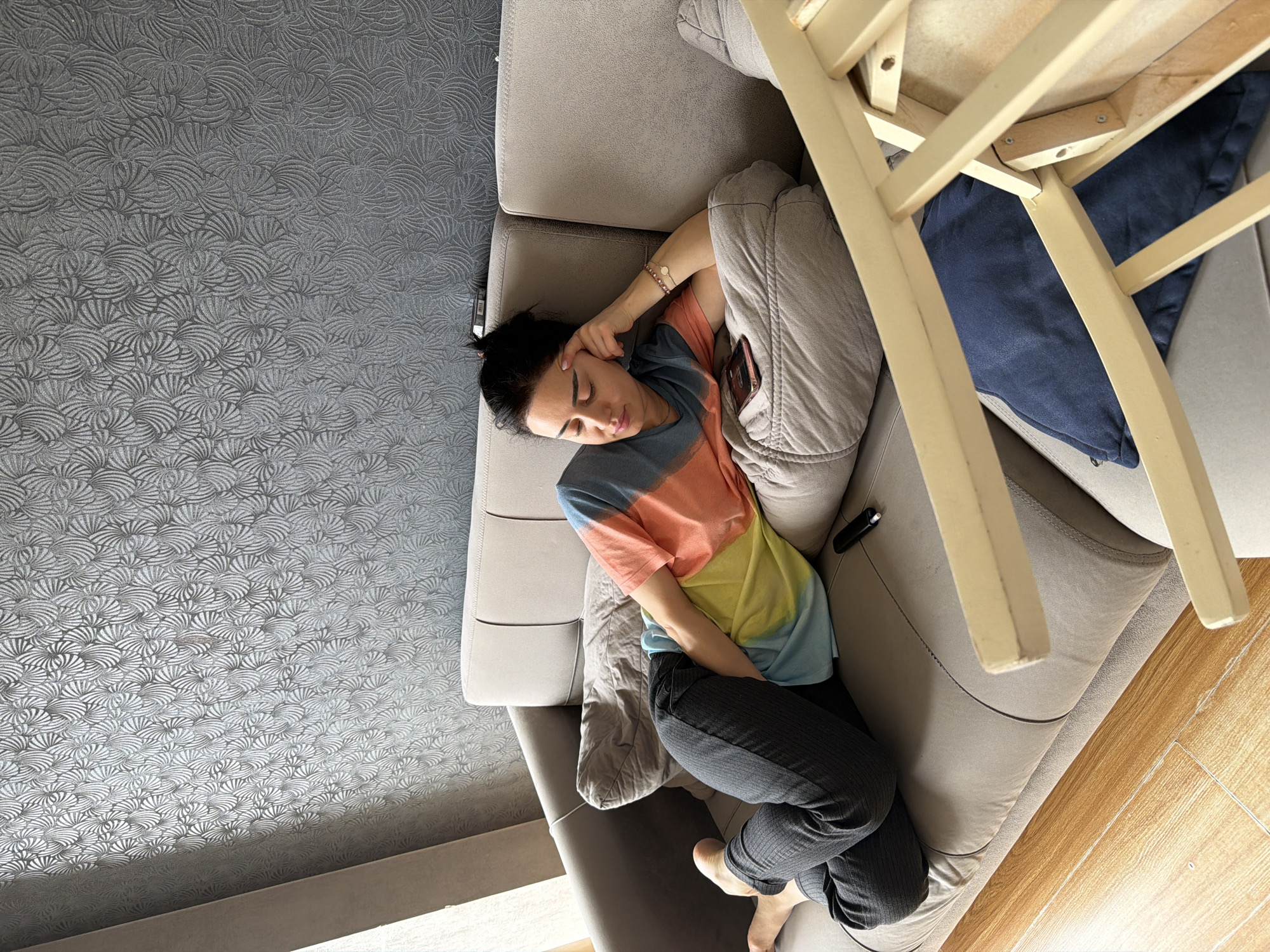 An overhead shot captures a person resting peacefully on a light grey sofa indoors in Tbilisi, Georgia. The subject, appearing to be a female, is lying on her left side, curled up with her eyes closed, suggesting sleep or deep relaxation. Her head is supported by her left hand, which is adorned with a beaded bracelet and a metallic wrist accessory. She is wearing a colorful tie-dye t-shirt featuring segments of blue, orange, and yellow, paired with dark, subtly patterned trousers. A smartphone is partially visible in a pocket or crease of her trousers.
A light wooden chair, possibly leaning or overturned, occupies the upper right portion of the frame, with a dark blue cushion visible beneath its structure. The flooring consists of two distinct materials: a dark grey carpet with an intricate, swirling, shell-like pattern covers the area to the left of the sofa, while a light wood-effect floor extends along the right side. A soft grey blanket or pillow is placed beside the person on the sofa. The scene is illuminated by natural light, indicating it is daytime, and evokes a tranquil, restful mood. No legible text is visible in the image.