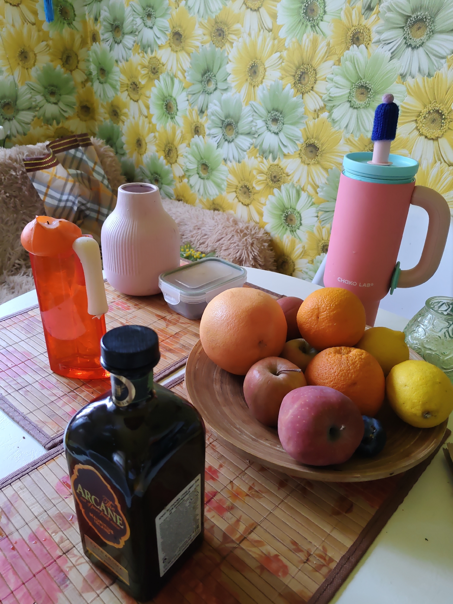 The image depicts a still life arrangement of various objects on a table, likely indoors. In the foreground, a dark glass bottle with a gold and black label reading "The Arcane" is prominently displayed. To the right, a wooden bowl contains a collection of fruits, including oranges, lemons, apples, and a dark plum. Behind the bowl, a pink insulated tumbler with a blue lid and a small knitted blue cover on its straw is visible. To the left of the tumbler, an orange plastic bottle holds a white candle. A light pink ribbed vase stands behind the orange bottle, and a small clear plastic container with a grey lid is situated between the vase and the fruit bowl. The background features a wall covered in floral wallpaper with yellow, green, and white daisy-like flowers. A plush brown object, possibly a stuffed animal or cushion, is partially visible on the left. The scene is lit by ambient lighting, casting soft shadows. No specific location in Rostov-on-Don, Russia, can be identified from the visual information provided.