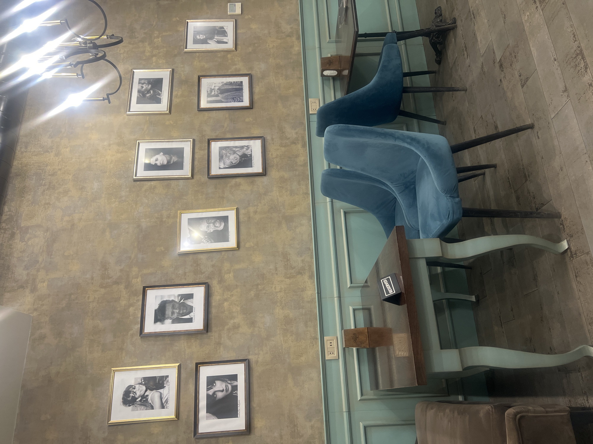 An interior view captures a stylish cafe or restaurant setting, likely located in Yerevan, Armenia. The left side of the image features a textured wall in shades of gold and brown, adorned with a collection of framed black and white portraits. These portraits depict various iconic figures, with visible names including Catherine Deneuve, Marilyn Monroe, Claudia Cardinale, Vincent Cassel, Isabella Rossellini, and "Antoine". A large, ornate black chandelier with multiple bright lights is prominent in the top left, casting a strong lens flare across the upper portion of the wall.
On the right, a seating area is visible with light blue/green paneled walls and a patterned tiled floor. Several elegant blue velvet chairs with dark legs are arranged around wooden tables that feature matching light blue/green legs. One table has a small black sign with the word "RESERVED" clearly visible in white text, and a small wooden box sits beside it. An electrical outlet is also visible on the paneled wall. A part of a brown upholstered chair is seen in the bottom right, suggesting additional seating. The establishment appears empty, contributing to a serene and inviting atmosphere.