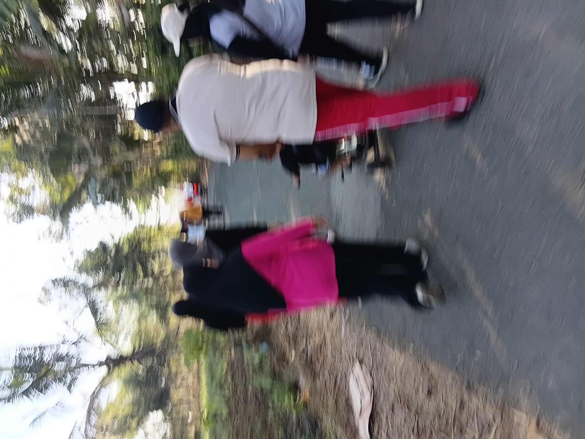 This is a motion-blurred image of several people walking on a paved road in Tanggul Wetan, Indonesia. The individuals are dressed in casual clothing. One person in the foreground is wearing a light-colored t-shirt and bright red pants. To their right, another person wears a light-colored t-shirt and dark pants, with a white cap on their head. A child is seated on a tricycle behind the person in the red pants. On the left side of the image, a person in a bright pink shirt and black pants is walking, and behind them, other figures are visible, including someone wearing a black headscarf. The background consists of trees and vegetation, suggesting an outdoor environment. The lighting indicates it is daytime.