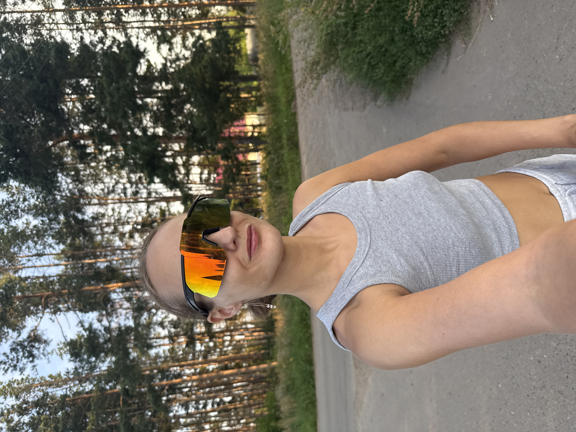 A young woman is captured in a selfie, outdoors in what appears to be a wooded area. She is wearing a grey tank top and reflective sunglasses. The sunglasses are a prominent feature, reflecting a vibrant orange and yellow sunset. Her hair is blonde and pulled back. She is wearing a delicate earring. The background suggests a path or road running alongside a dense pine forest. The overall lighting indicates it is late afternoon or early evening, with the warm hues of sunset visible in the sky and reflecting in her eyewear. The scene evokes a sense of casual outdoor activity, possibly a walk or run. No text is visible in the image. The location context provided is Подшивалово, Russia.