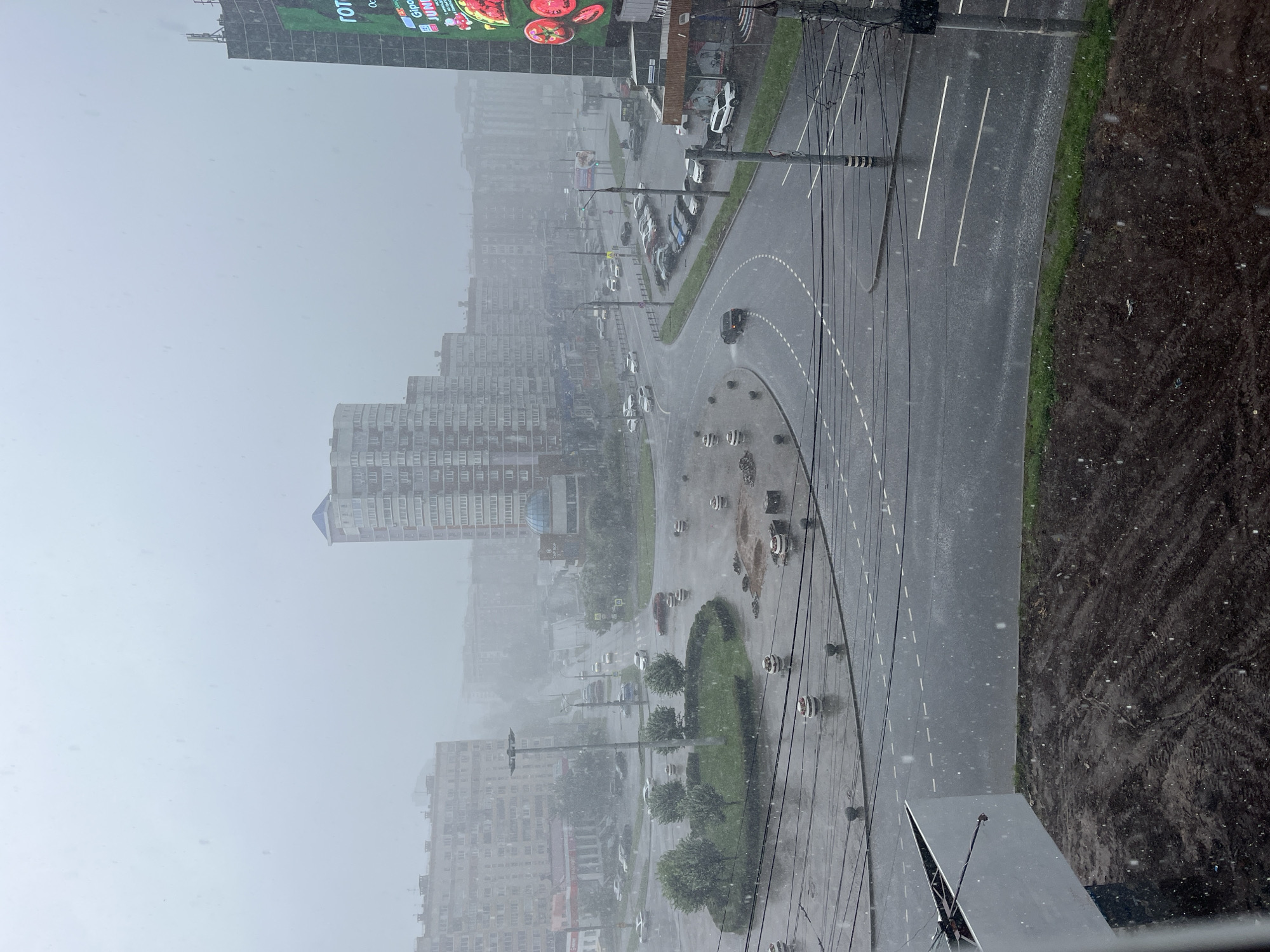 The image captures a rainy urban scene in Izhevsk, Russia, likely from a high vantage point, overlooking a street intersection and a circular green space with trees. The sky is overcast and a heavy downpour is evident, with raindrops visible throughout the frame and on the wet asphalt. Several cars are driving on the roads, some appearing to be stopped at the intersection. Buildings of varying heights, characteristic of Soviet-era urban planning, line the background, and a large digital billboard is visible on the right side of the frame. The foreground shows a construction area with dark soil and some fencing. The overall impression is one of a somber, wet day in a typical Russian city.