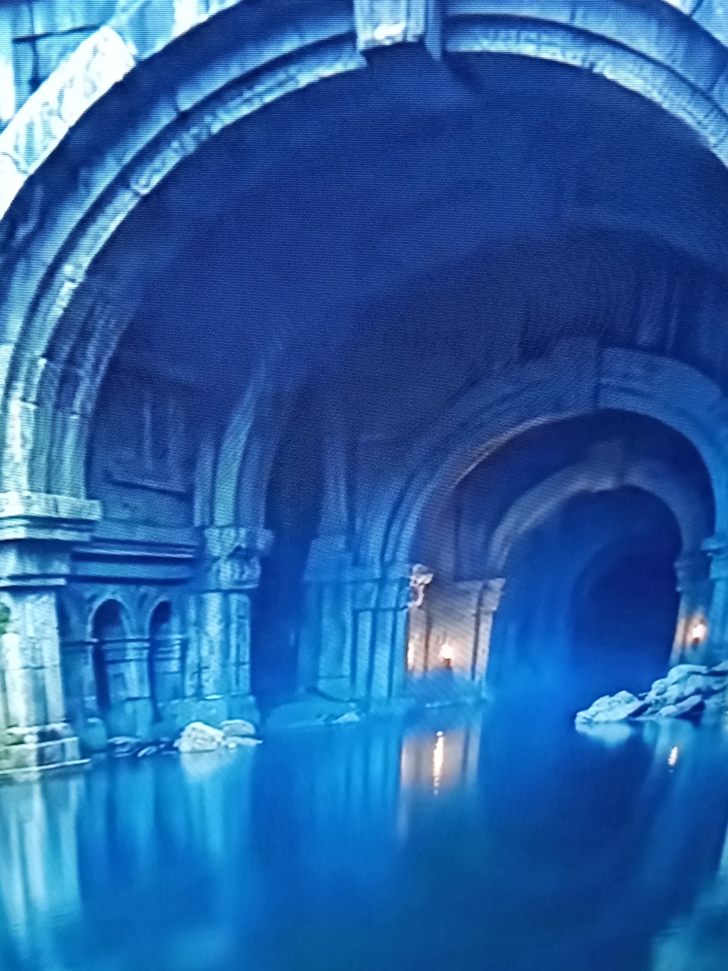 The image depicts an interior view of a large, ancient-looking stone structure with multiple arched passages. Water fills the floor of this structure, reflecting the dim light from torches or lamps visible within the passages. The architecture features stone columns, arches, and carved details, suggesting a historical or fantasy setting. There are no people visible in the scene. The environment is dark, with a dominant blue-toned lighting, creating a mysterious and somewhat somber atmosphere. The scene is not identifiable as being located in the city of Brno, Czechia.