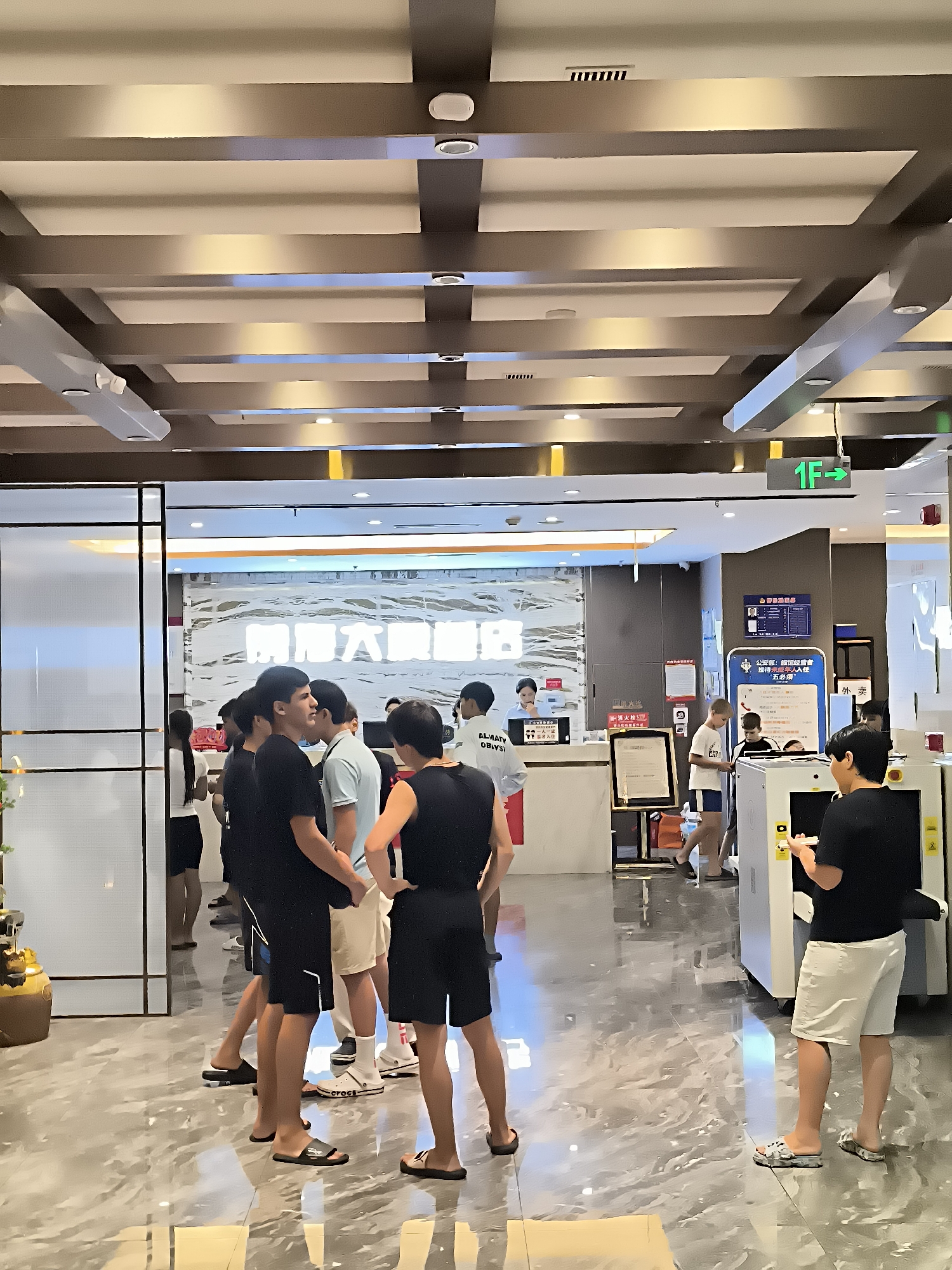 The image captures a scene inside a brightly lit building with a modern ceiling design featuring exposed beams and recessed lighting. In the foreground, a group of people, mostly young men, are gathered. Some are standing and talking, while others appear to be waiting in line. One individual is seen interacting with a white electronic device, possibly a scanner or kiosk. In the background, there is a reception desk area with a person behind it, and several signs are visible on the walls. One sign prominently displays "1F" with a green arrow pointing right, indicating the first floor and a direction. The flooring is a polished marble or similar material. The overall impression is that of a public service building or a commercial establishment, potentially a government office or a service center, in Ürümqi, China.
