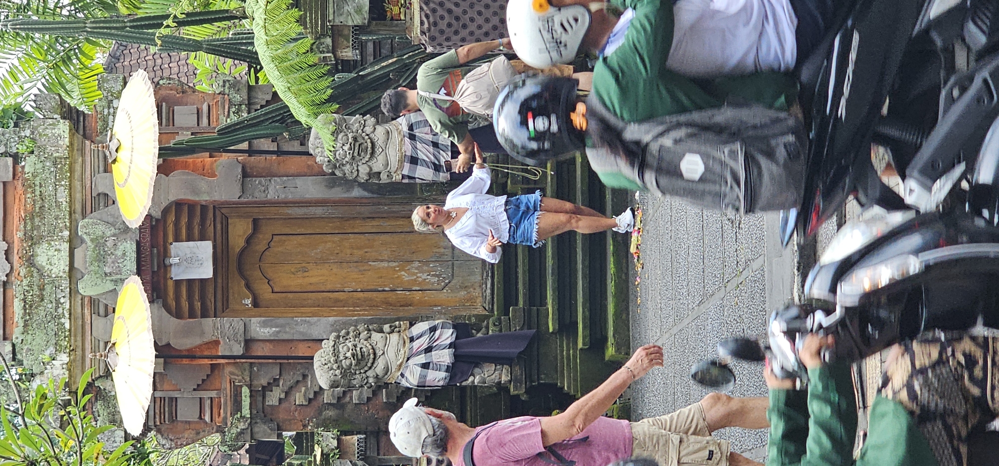 The image, taken in the Ubud District of Indonesia, features a wooden temple entrance flanked by stone statues draped in checkered fabric. Two yellow ceremonial umbrellas adorn the temple's facade. A woman in a white blouse and denim shorts stands near the temple entrance. Several other people are present, including a man in a pink shirt and khaki shorts, and several people wearing helmets and riding motorcycles. The ground is stone and covered with offerings, and lush greenery surrounds the temple structure.