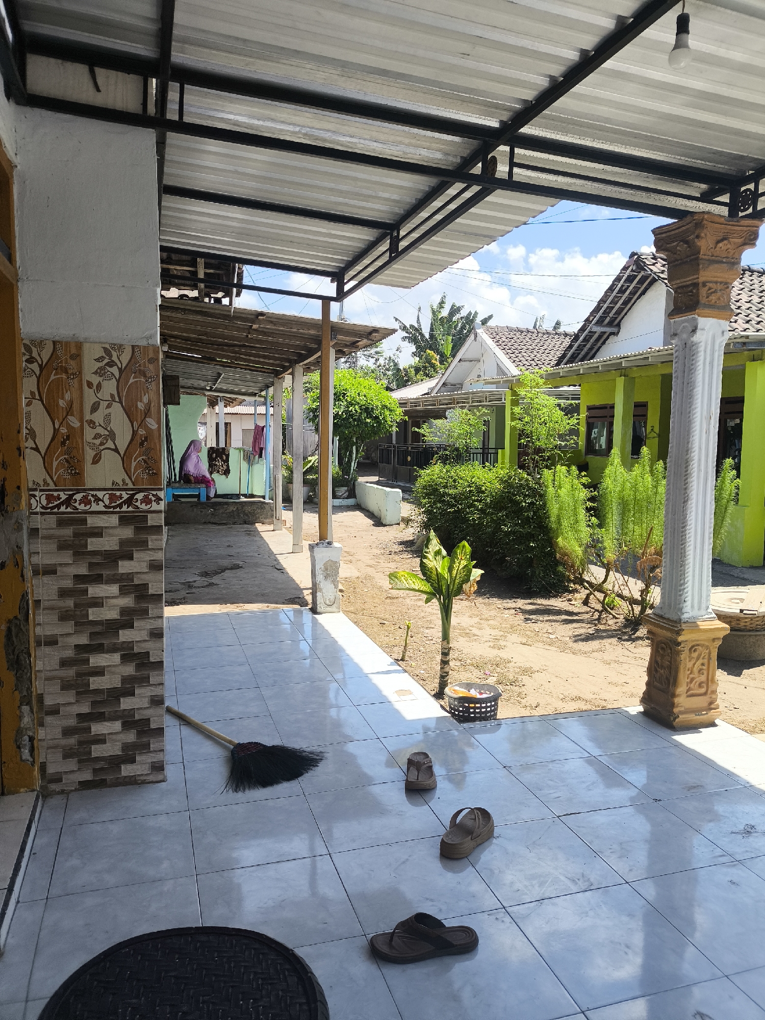 The image depicts a view from a covered porch looking out into a residential yard. The porch floor is tiled with large, light-colored square tiles, and a broom leans against a tiled pillar. In the yard, there are several pairs of sandals scattered on the ground. Beyond the yard, there are brightly painted green houses with tiled roofs, typical of residential architecture in Grati, Indonesia. Lush green bushes and trees are visible, and the sky is a clear blue with some scattered clouds. The scene suggests a casual, everyday setting, possibly a home environment in a rural or semi-rural area of Grati.