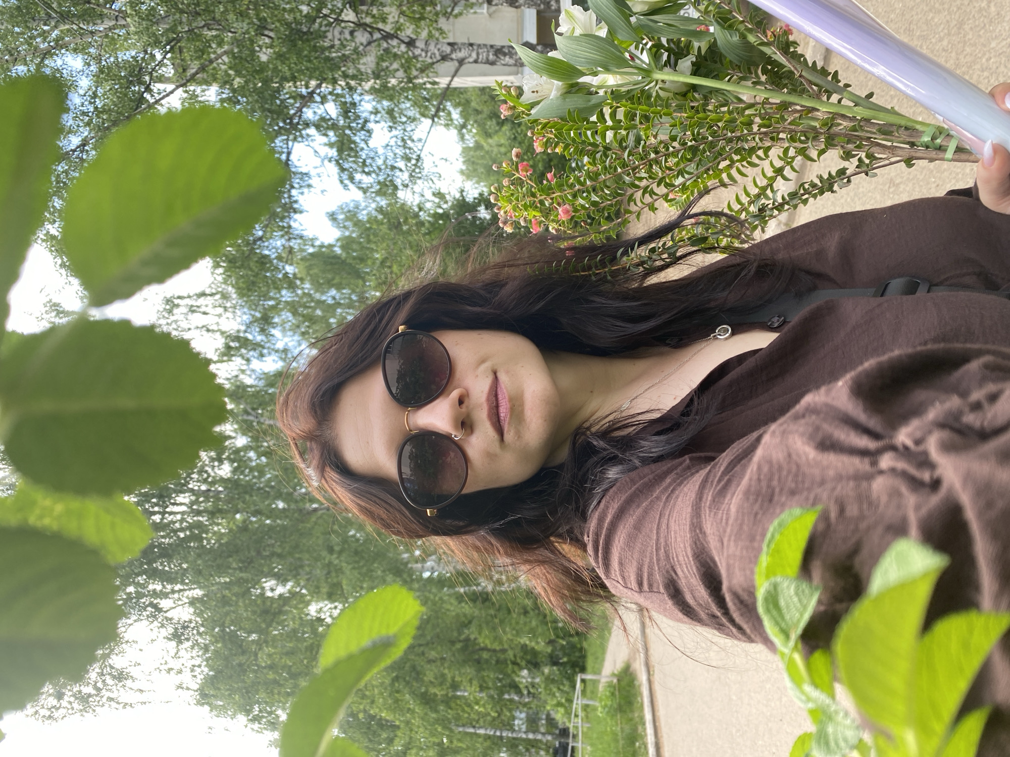 A young woman with dark hair and sunglasses holds a bouquet of white lilies and green foliage. She wears a brown shirt and a silver necklace. The background shows trees and a sidewalk, suggesting an outdoor setting, possibly a park or street in the city. The location is given as Chaikovsky, Russia. The photograph seems to be taken at a low angle with some leaves in the foreground, giving a sense of depth to the image.