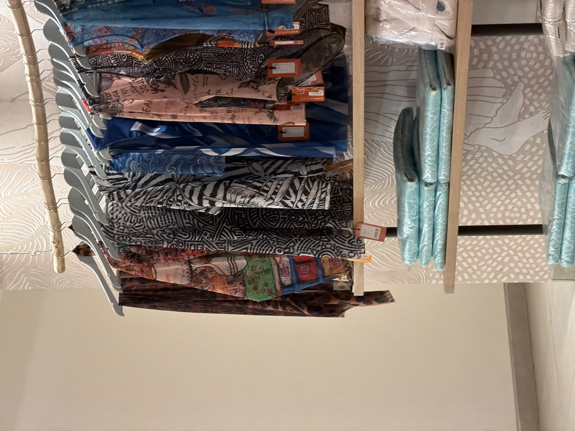 This image shows a display of clothing items, likely in a retail setting in Boreti, Montenegro. The subjects are various garments, predominantly tops and scarves, folded and hung on hangers. The clothes exhibit a variety of bold patterns, including tribal prints, abstract designs, and a leopard print. Some of the fabrics appear lightweight and sheer, suggesting they might be summer wear.
On the right side of the image, there are several items wrapped in translucent blue plastic, possibly representing another style of apparel or accessories. The display is organized on wooden shelves and hangers, with a textured, pale background featuring a repetitive pattern of white lines and dots.
Several small orange tags are attached to some of the garments, indicating pricing or branding information. One tag clearly shows the text "RODA SOLEIL". The lighting is bright and even, suggesting an indoor environment, possibly a boutique or a section of a larger store. No people are visible in the image, and there are no overt cues about the time of day or weather. The overall impression is one of a colorful and patterned clothing display.