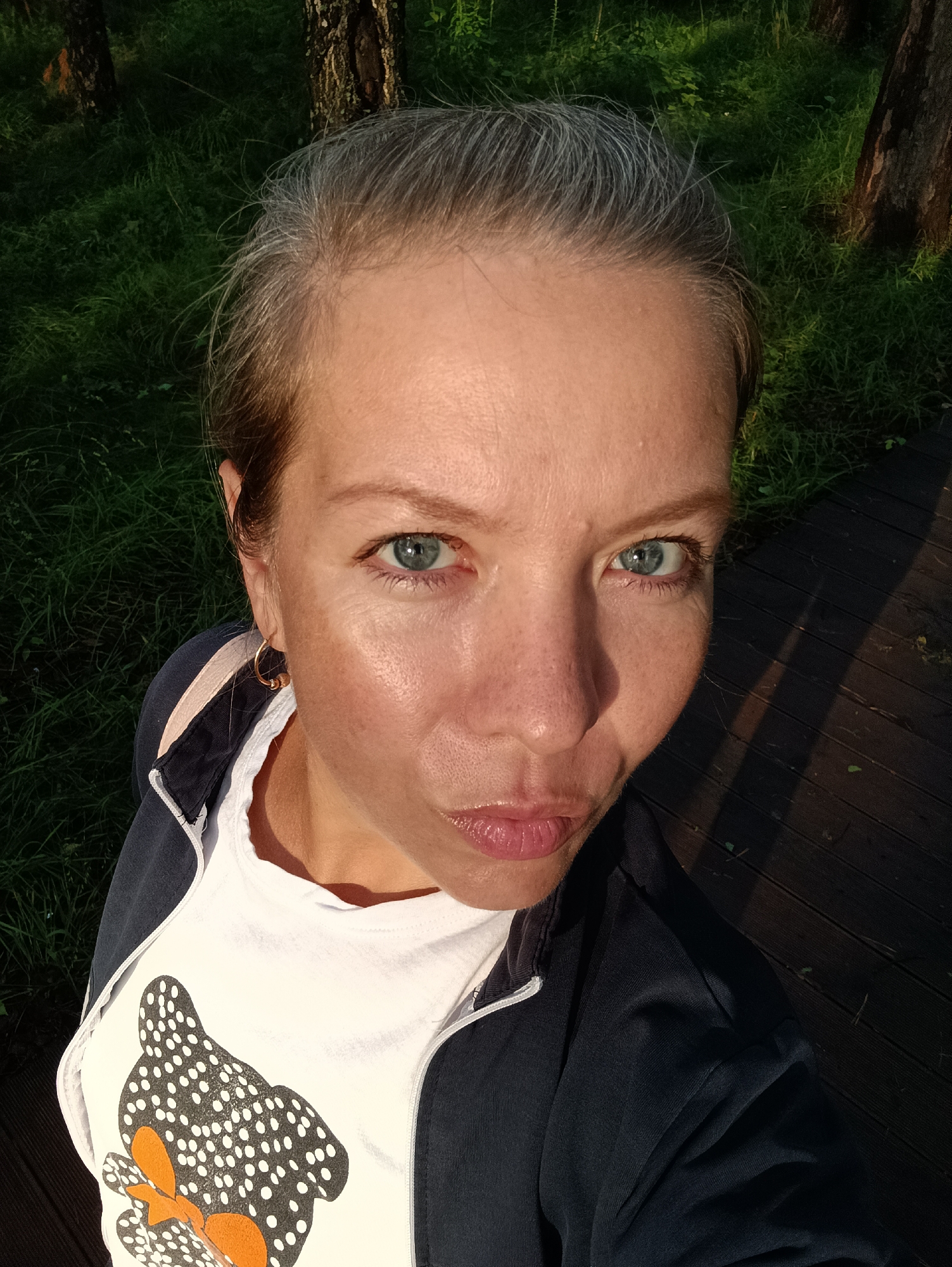 A high-angle, outdoor, close-up selfie shows a fair-skinned woman with grey hair pulled back and piercing blue eyes. She is wearing a white t-shirt with a black and white spotted design of a cartoonish animal and a dark blue jacket. The woman has a subtle pout and is looking directly at the camera. Her left earlobe is adorned with a small, gold hoop earring.
The background is a blur of natural elements. To the left and top of the frame, lush green grass and trees suggest a park or wooded area. To the right, a dark wooden boardwalk is visible, casting distinct shadows that indicate it is late afternoon or early evening. The overall setting appears to be a public outdoor space in a city. Given the request to include the city of Chaikovsky, Russia, this scene is likely located in a park within that city, possibly near a recreational area or walking path.