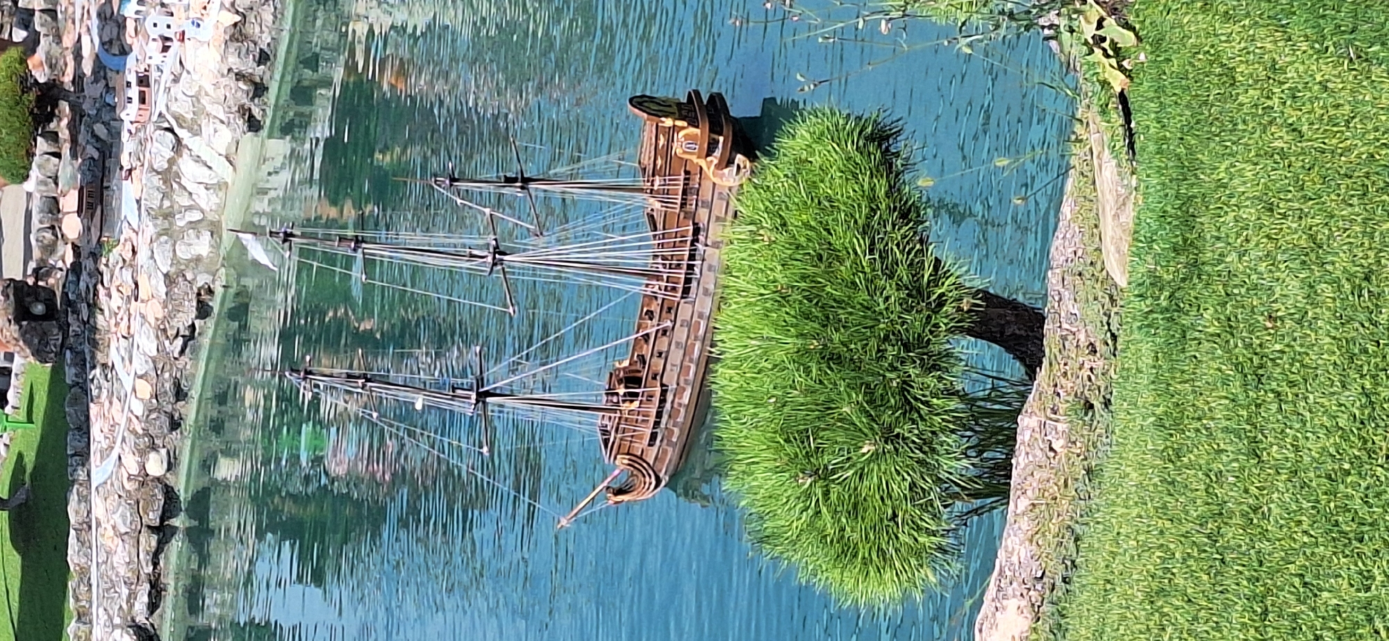 The image depicts a miniature sailing ship model floating on a body of water. The ship is brown with multiple masts and rigging. In the foreground, a lush green tree with a thick trunk is partially submerged in the water, with its foliage reaching out over the surface. The water is a clear turquoise color, reflecting the sky and surroundings. The background shows a rocky embankment with miniature buildings and landscaping, suggesting this is a diorama or a model village. The scene appears to be outdoors, under daylight. This scene is located at Italia in Miniatura, a miniature park in Viserbella, a frazione of Rimini, Italy.