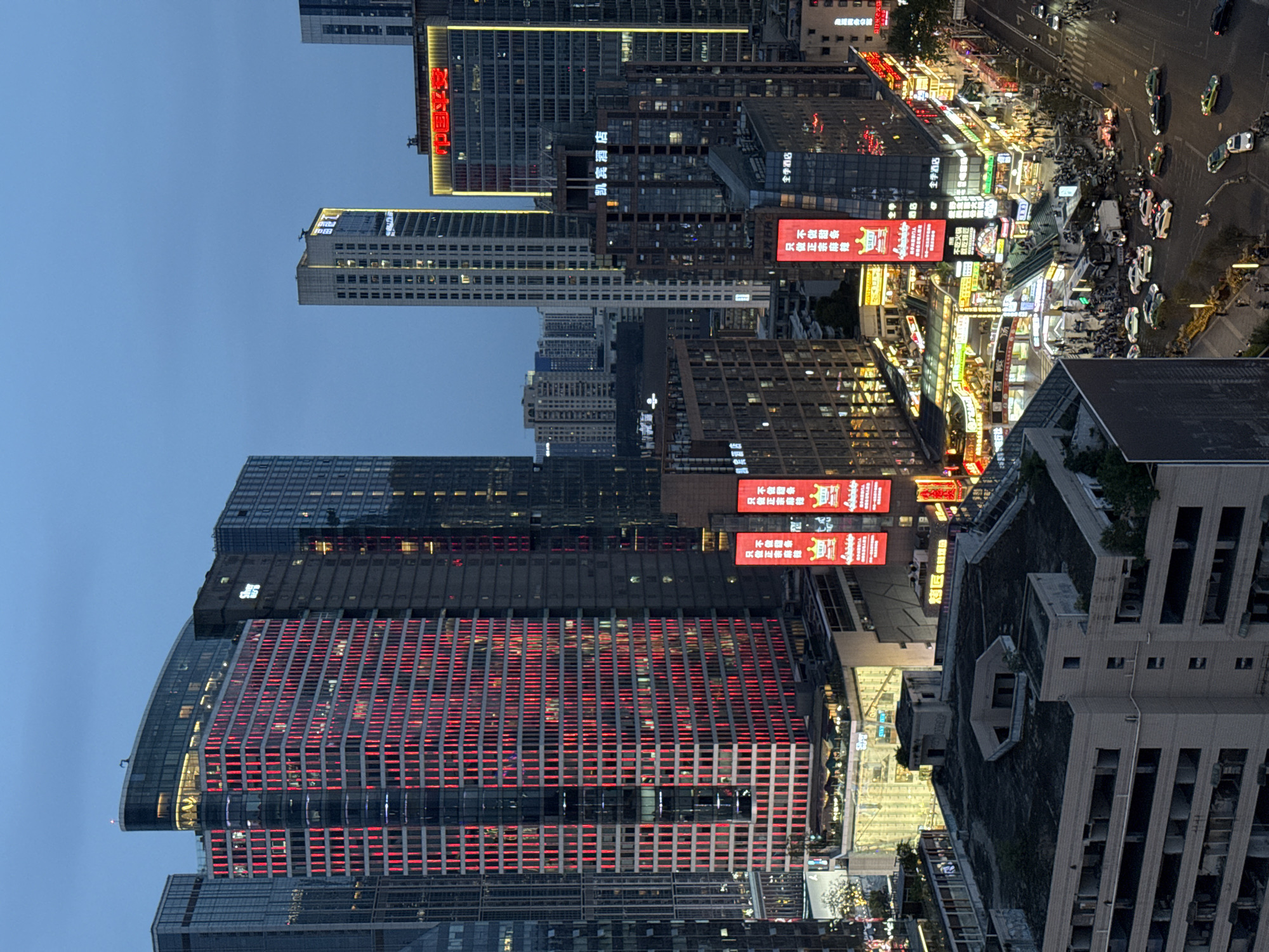 The image depicts an urban cityscape at dusk, likely located in the Jinjiang District of China. Prominent architectural features include several skyscrapers, one of which is notably illuminated with red horizontal bands across its facade. Brightly lit commercial streets and buildings contrast with the fading natural light. The streets below show moving vehicles and what appear to be pedestrians, indicating an active urban environment. Chinese signage is visible on various buildings and billboards, adding to the location's visual context.