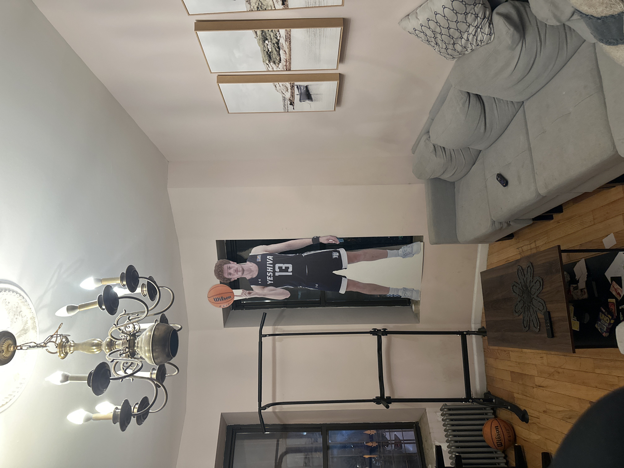 The image depicts an interior scene. On the left, a chandelier hangs from the ceiling, featuring multiple curved arms holding light bulbs. Moving to the right, a series of framed landscape prints adorn the wall above a poster of a basketball player wearing a jersey with "YESHIVA 13" printed on it, holding a Wilson basketball. Below the poster, a black garment rack stands near a window, and a radiator is visible next to a basketball on the floor. In the upper right corner, a section of a grey sectional couch is visible, along with a patterned pillow. A dark wooden coffee table is positioned in front of the couch. The scene appears to be indoors, possibly a residence in New York City, United States of America, due to the presence of items such as a radiator, chandelier, and framed artwork, indicative of a domestic setting.