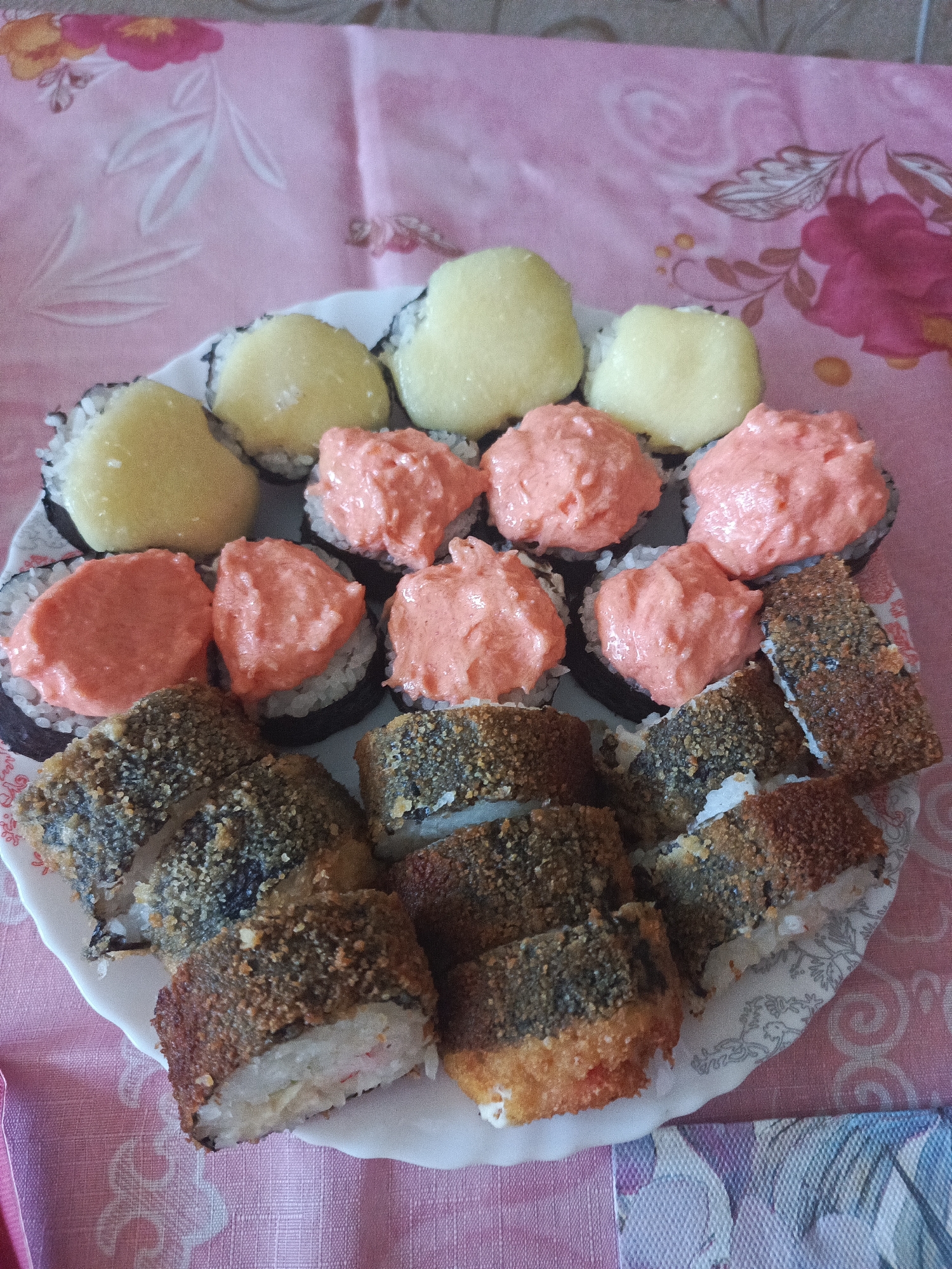 A white, circular plate holds a variety of sushi rolls arranged in two concentric circles. The inner circle features several rolls topped with a pale yellow, creamy substance. The outer circle contains rolls topped with a pink, creamy mixture, as well as several rolls that are breaded and deep-fried, showing a golden-brown crust. The plate sits on a pink tablecloth with a floral pattern. The lighting suggests an indoor setting.