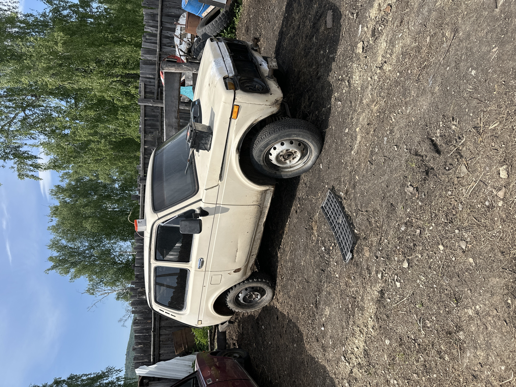 The image depicts a white Lada Niva parked on a dirt surface. The location is an outdoor space, possibly a junkyard or rural area in an unknown city. The car appears to be in a state of disrepair, with some missing parts. A wooden fence runs along one side of the frame, and a tree with green leaves is visible behind the fence. A metal grate sits on the ground near the vehicle. The sky is visible with blue coloring, suggesting it's daytime.