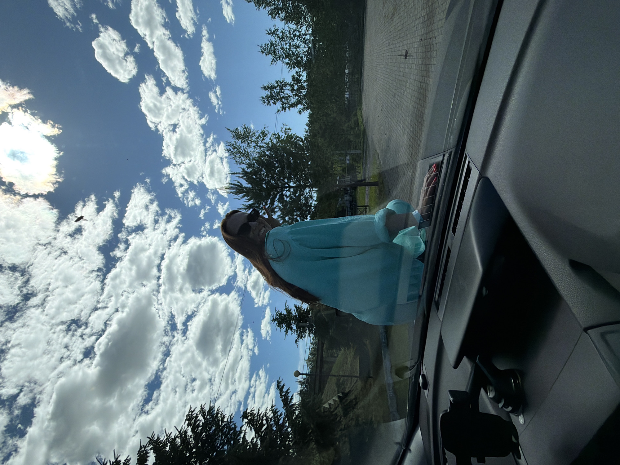 An overhead, wide-angle shot looks down at a woman from the perspective of inside a car. The woman is sitting on the hood of the car, facing away from the camera and looking over her shoulder. She has long, brown hair and is wearing sunglasses and a light blue and white striped long-sleeved shirt. Her fingernails are painted red. The car's dashboard is visible in the foreground, with a phone mount attached to the windshield.
The background shows a bright blue sky filled with white, puffy clouds. The sun is visible in the upper right corner, with a halo effect. Trees and a light-colored paved area are visible in the middle ground. A bridge and more trees are in the distance. The overall scene suggests an outdoor setting, possibly a parking lot or roadside. The specific location is not identifiable from the image itself, and any mention of Birobidzhan, Russia, would be an assumption not supported by visual evidence.