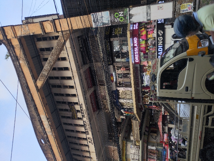 The image depicts a multi-story commercial building identified as "J.D VASTRA MAHAL" located on Market Road in Ernakulam, India, under daylight conditions. The building exhibits a weathered concrete facade with multiple balconies and metal railings across at least four visible levels. Numerous commercial establishments are present within, including "PERFECT COLLECTION," "DRESSLAND" (displaying colorful children's garments on mannequins), and "KARNITAB'S." Overhead utility cables crisscross the frame in front of the building. In the mid-ground, a light-colored pickup truck with a yellow front bumper is positioned, facing right, partially obscuring the lower levels of the building. To the right, a person wearing a blue helmet and a light-colored shirt is partially visible, likely on a two-wheeler. In the left background, behind the truck, several two-wheelers are parked, and multiple individuals are present near them and on the building's balconies, suggesting typical street activity in a commercial area. A large vertical sign on the far right displays text including "STAY TUNED SO ON."