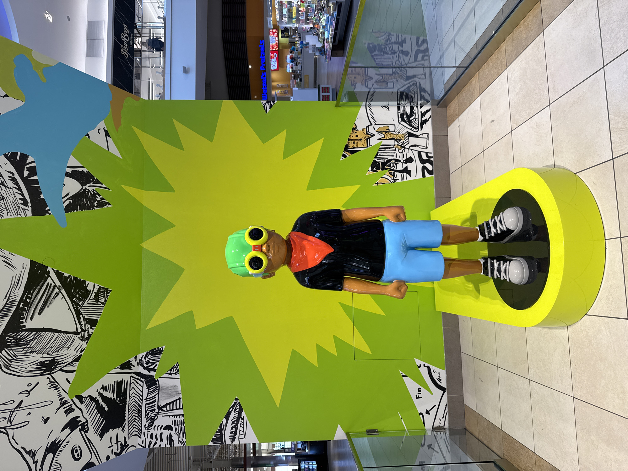 This media file presents a vibrant, colorful statue of a child or young person displayed within a mall environment in Rosemont, USA. The image is oriented with the statue standing vertically, although the original photo appears to have been taken in landscape and rotated.

The primary subject is a statue depicted as a light-skinned individual with dark brown skin tone, standing upright. They wear a bright green helmet with large yellow goggles, an orange bandana around the neck, a black short-sleeved shirt, light blue shorts, and black sneakers with white laces. The statue is positioned on a bright yellow, circular base.

Behind the statue is a wall designed in a comic book style. A prominent feature is a large, bright green "explosion" or "starburst" shape that emanates from behind the statue. Parts of the wall around this green shape, particularly to the left and right, are covered with black and white comic book-style illustrations featuring dynamic lines, faces, and abstract elements. A segment of light blue and brown color, resembling a map or stylized landmass, is visible on the upper left portion of the wall.

The setting is clearly an indoor shopping mall. In the background, beyond the display wall and glass railings, other stores are visible. Text on a store sign reads "Kiehl's Since 1851". Another sign further in the distance displays "Wetzels Pretzels". The floor is tiled in a light, neutral color. The overall atmosphere is brightly lit, typical of a mall during its operating hours. The artistic display evokes a sense of fun, adventure, and youthfulness. Visible fragmented text on the comic-style wall includes "Fin" and other illegible comic book sound effects or dialogue bubbles. The sneakers on the statue appear to have "KKKII" printed on them, likely part of a brand name.
