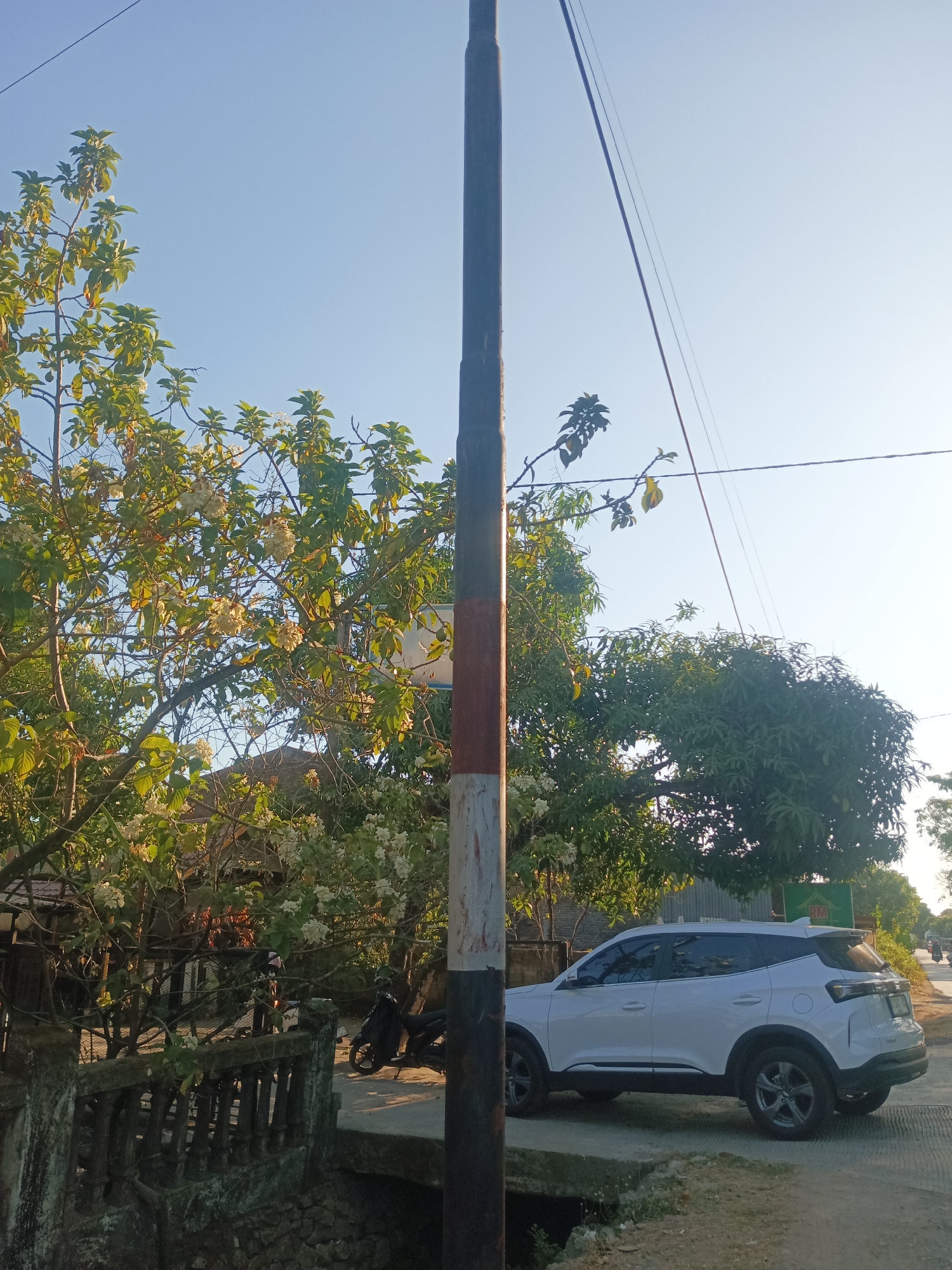 The image captures a street scene in Bowong Cindea, Indonesia, bathed in the warm light of a clear, sunny day. A tall, striped utility pole stands prominently in the foreground, a dark brown, white, and red banded post. To its right, a white SUV is parked on a paved surface, its rear facing the viewer and its front angled slightly towards the right edge of the frame. The vehicle appears to be a modern crossover with tinted windows. Behind the SUV, a lush green tree with dense foliage dominates the middle ground. To the left of the pole, a flowering tree with white blossoms and vibrant green leaves is in full bloom. A stone balustrade runs horizontally across the lower left of the image, bordering a dark, unseen area. Further in the background, a building with a weathered roof is visible amongst the trees. A black motorcycle is parked near the balustrade. The sky above is a serene, pale blue. The overall impression is one of a quiet, sun-drenched day in a semi-rural or suburban setting in Indonesia.