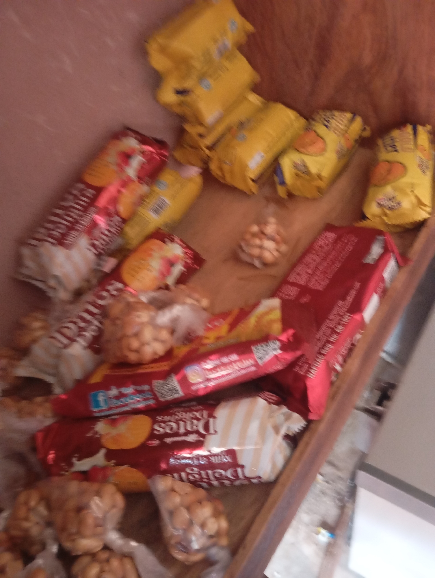 The image depicts a close-up, angled view of a collection of packaged snacks and small, round food items, likely nuts or candies, arranged on a wooden surface. The items are predominantly in yellow and red packaging. Some of the yellow packages appear to be larger, multi-snack bags, while others are smaller, individual servings. The red packages are sleeker, and some feature images of fruit, possibly dates, and text in a non-English script. Several clear plastic bags contain clusters of the small, round food items. The lighting is dim and uneven, suggesting an indoor environment with limited light sources. The overall impression is of a display or storage of food items, possibly in a market stall or a shop. There are no people or animals visible in the image, and no specific context regarding the city of Jalingo, Nigeria, is evident from the visual information alone.