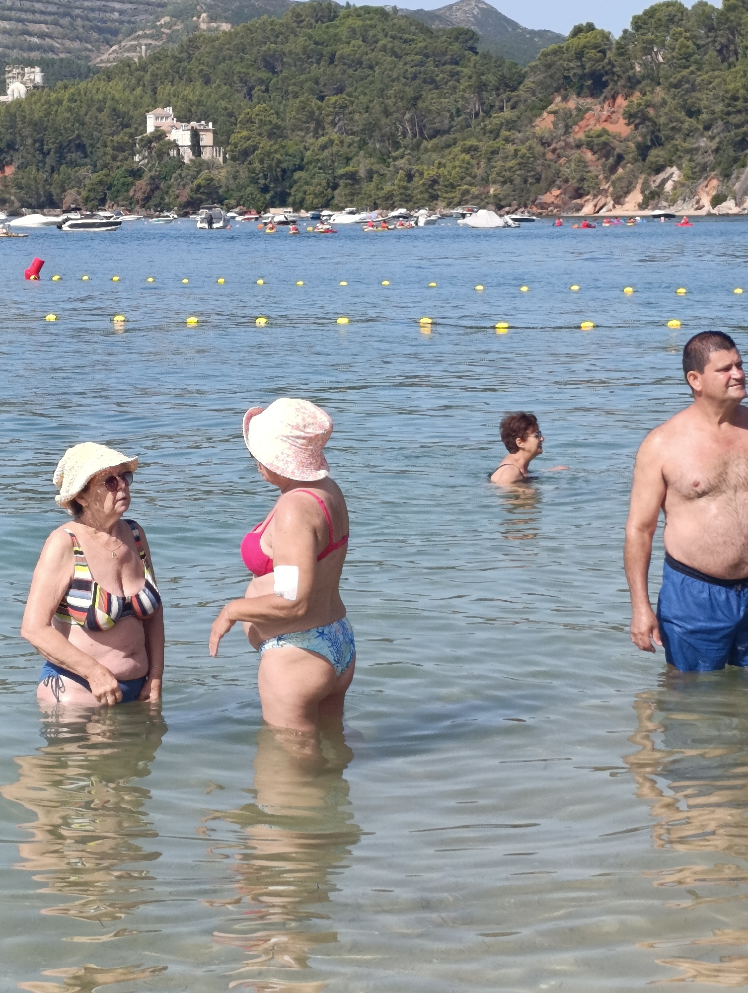 The image depicts a sunlit outdoor scene of people in the shallow waters of a beach, possibly in Setúbal, Portugal, given the presence of boats and a hilly, forested coastline in the background. In the foreground, three individuals are partially submerged in the clear blue water. On the left, an older woman wearing a striped bikini and a straw hat is facing a second older woman who is wearing a pink bikini top and patterned bottoms. The second woman has a bandage on her left arm. To the right of the frame, a man in blue swim shorts stands facing away from the viewer, looking to the right. Further in the background, another woman is swimming. The water is calm, with some ripples and reflections visible. Yellow buoys are dotted across the water, marking a designated area. Boats are visible in the middle ground, with a white building situated on a tree-covered hillside behind them, suggesting a coastal town or resort. The sky is clear and blue.
