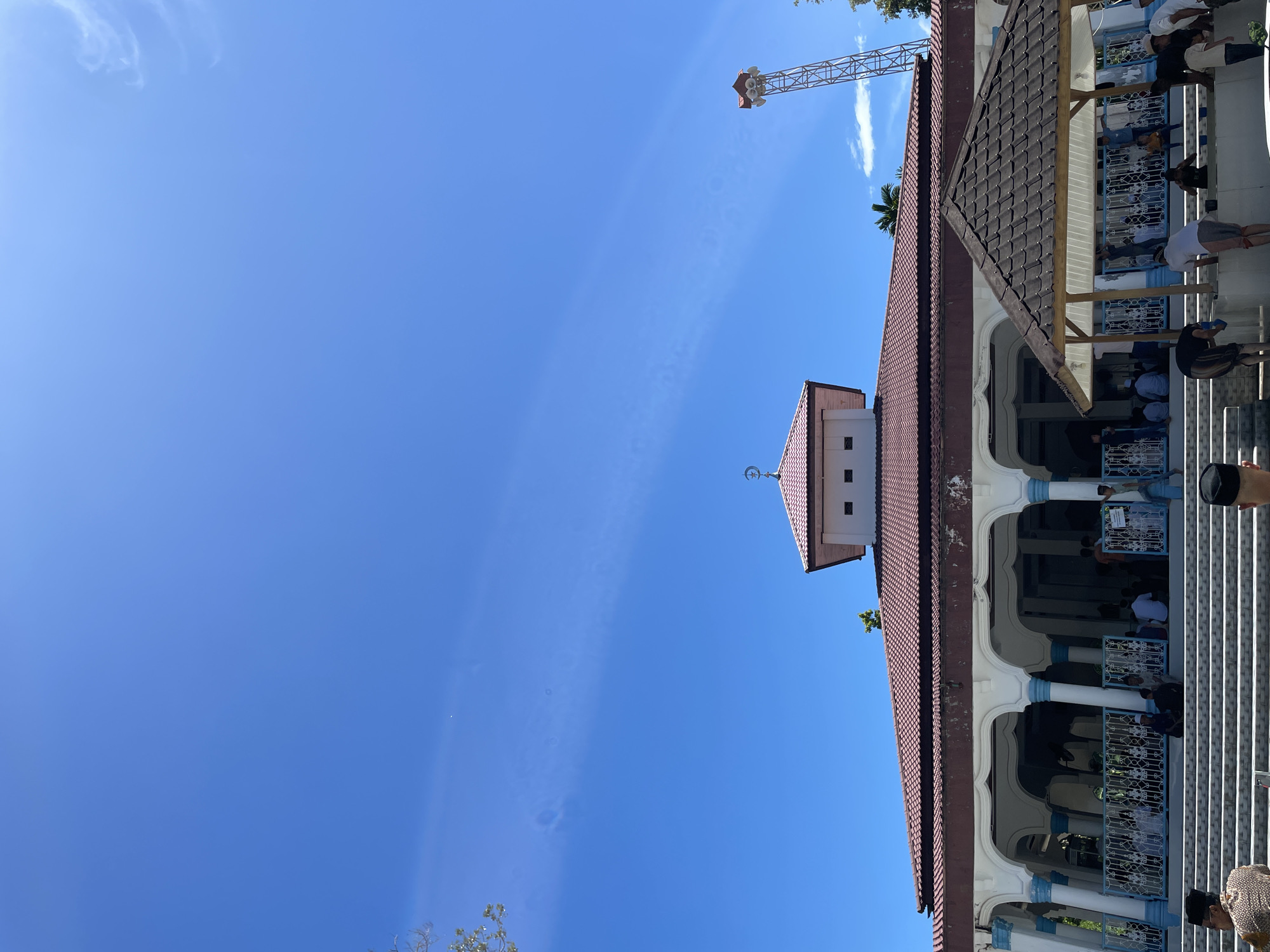 The image depicts a building with a red-tiled roof and white arched openings, possibly a mosque or a religious structure, under a clear blue sky. People are visible inside the building, some appearing to be engaged in prayer or other religious activities. A metal tower structure with speakers is also visible against the sky in the distance. The structure is located in or near Pasar Paloh, Indonesia, based on the location context.