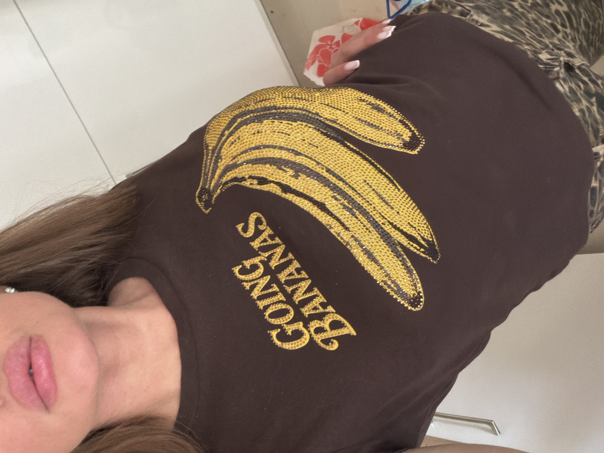 A woman wears a brown t-shirt with a yellow banana graphic and the words "GOING BANANAS." She has light skin, light pink lips, and long brown hair. She is wearing a leopard-print garment. The background is a white surface. The location is unknown and cannot be determined from the image.