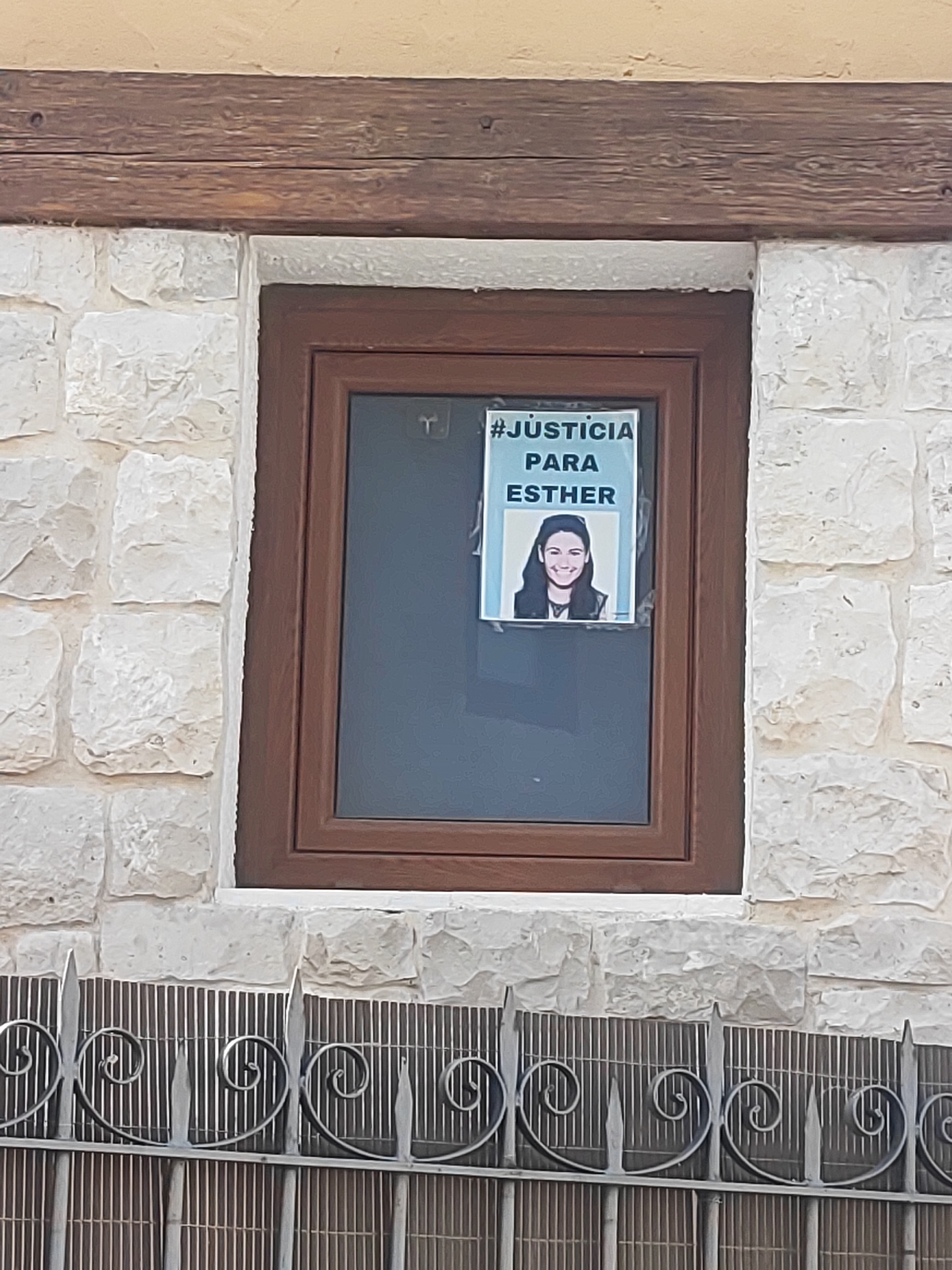 A poster reading "#JUSTICIA PARA ESTHER" (Justice for Esther) with a picture of a young woman is taped to the window of a house. The house has a stone facade and a wooden beam above the window. A metal fence with decorative swirls is in front of the house. The image was taken in the city of Traspinedo, Spain.