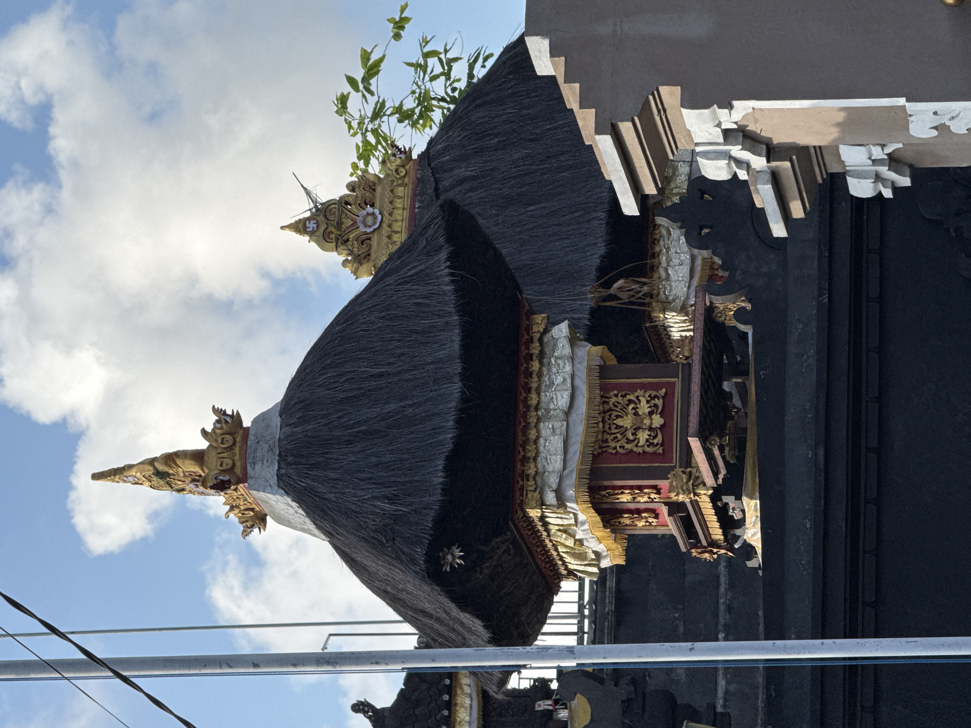 A close-up, low-angle outdoor photograph captures intricate Balinese temple architecture against a blue sky with scattered white clouds. In the foreground, a tiered, black thatched roof structure is adorned with a gilded, ornate finial featuring mythological figures. Below the thatched roof, a smaller red and gold structure is visible, decorated with intricate carvings and gilded fabric draped around its edges. To the right, a portion of another similar temple structure with a thatched roof is visible, also topped with a golden ornamental element. A green leafy branch extends into the frame from the right, reaching towards the temple structures. In the left of the frame, a white pole with electrical wires running horizontally is present. The foreground also shows dark, textured stone walls and architectural details of what appears to be a larger structure, possibly part of the temple complex, with layered, decorative moldings. The scene depicts a religious or cultural site, likely within the city of Kuta, Indonesia, known for its Hindu temples and traditional Balinese architecture.