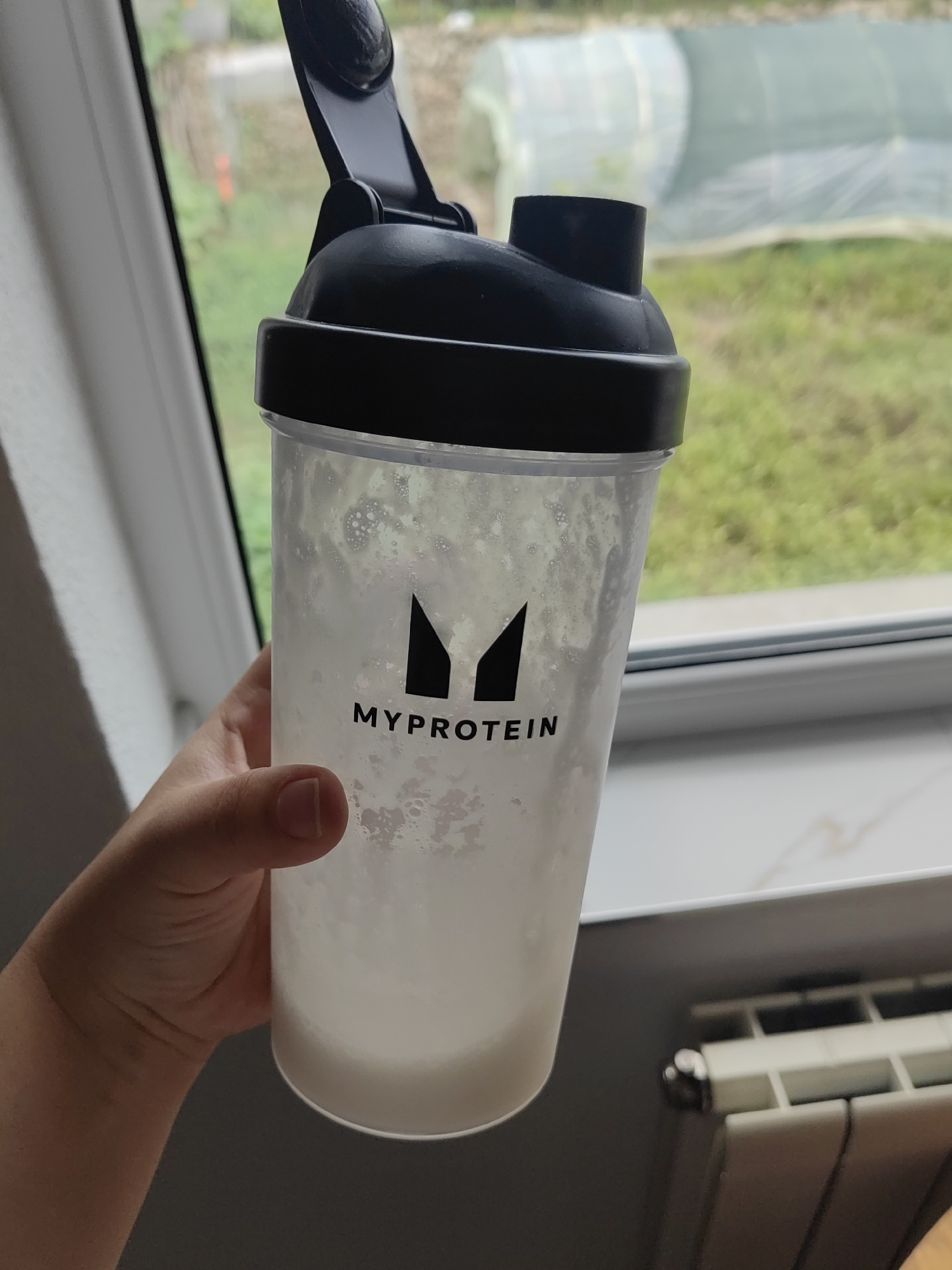 In Djerovo, Bulgaria, a person holds a transparent shaker bottle filled with a frothy, off-white liquid. The bottle has a black lid and a black "MYPROTEIN" logo. The background shows a blurry view through a window, with green foliage and what appears to be a greenhouse structure. A white windowsill and a section of a radiator are also visible. The lighting suggests it is daytime, possibly overcast.