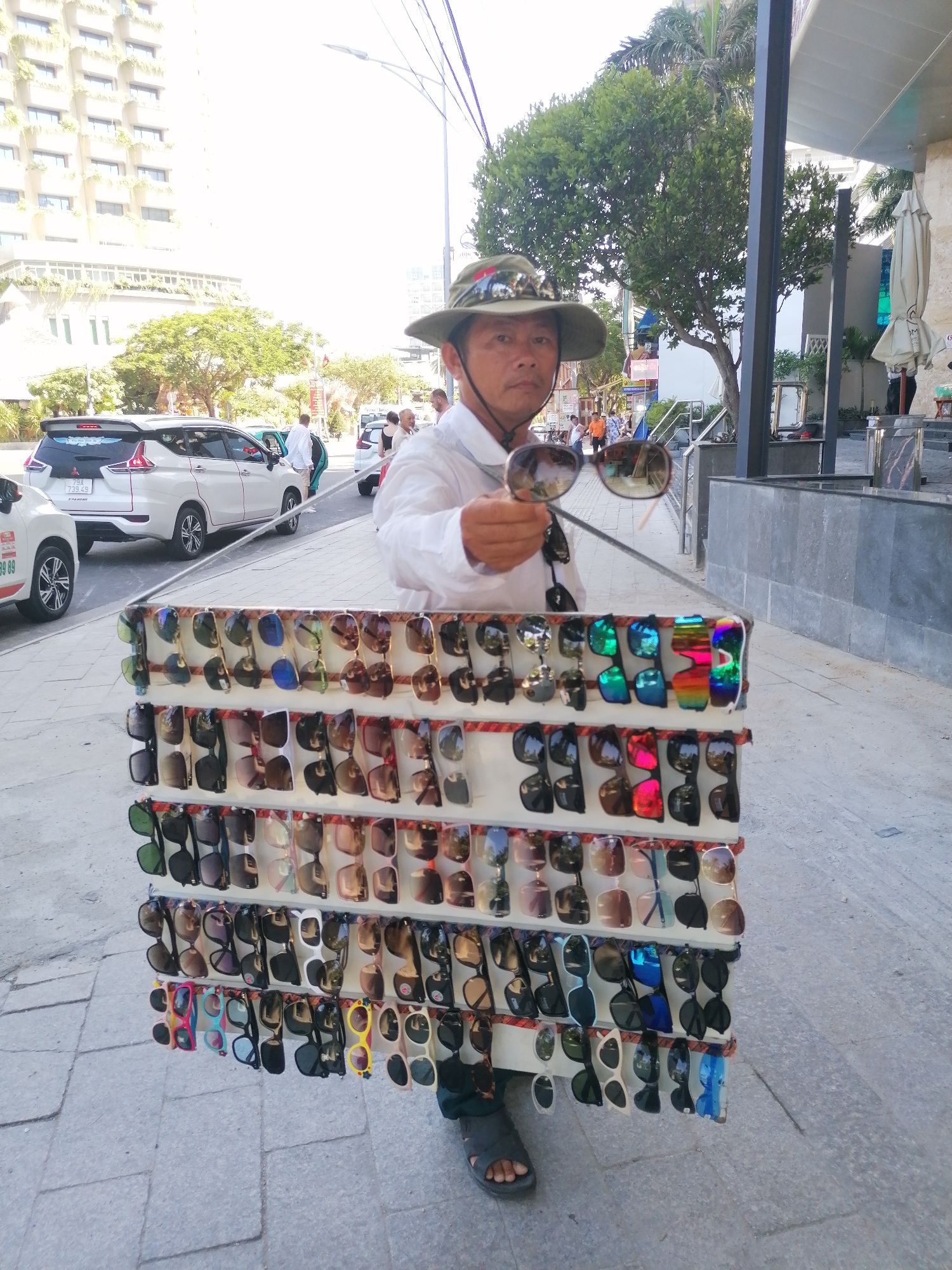 A street vendor stands in Nha Trang Ward, Vietnam, displaying a large selection of sunglasses. The vendor, a middle-aged man wearing a hat and white shirt, holds a pair of sunglasses in his right hand. The sunglasses are displayed on a white stand that he carries. In the background, there are cars, pedestrians, buildings, and trees.