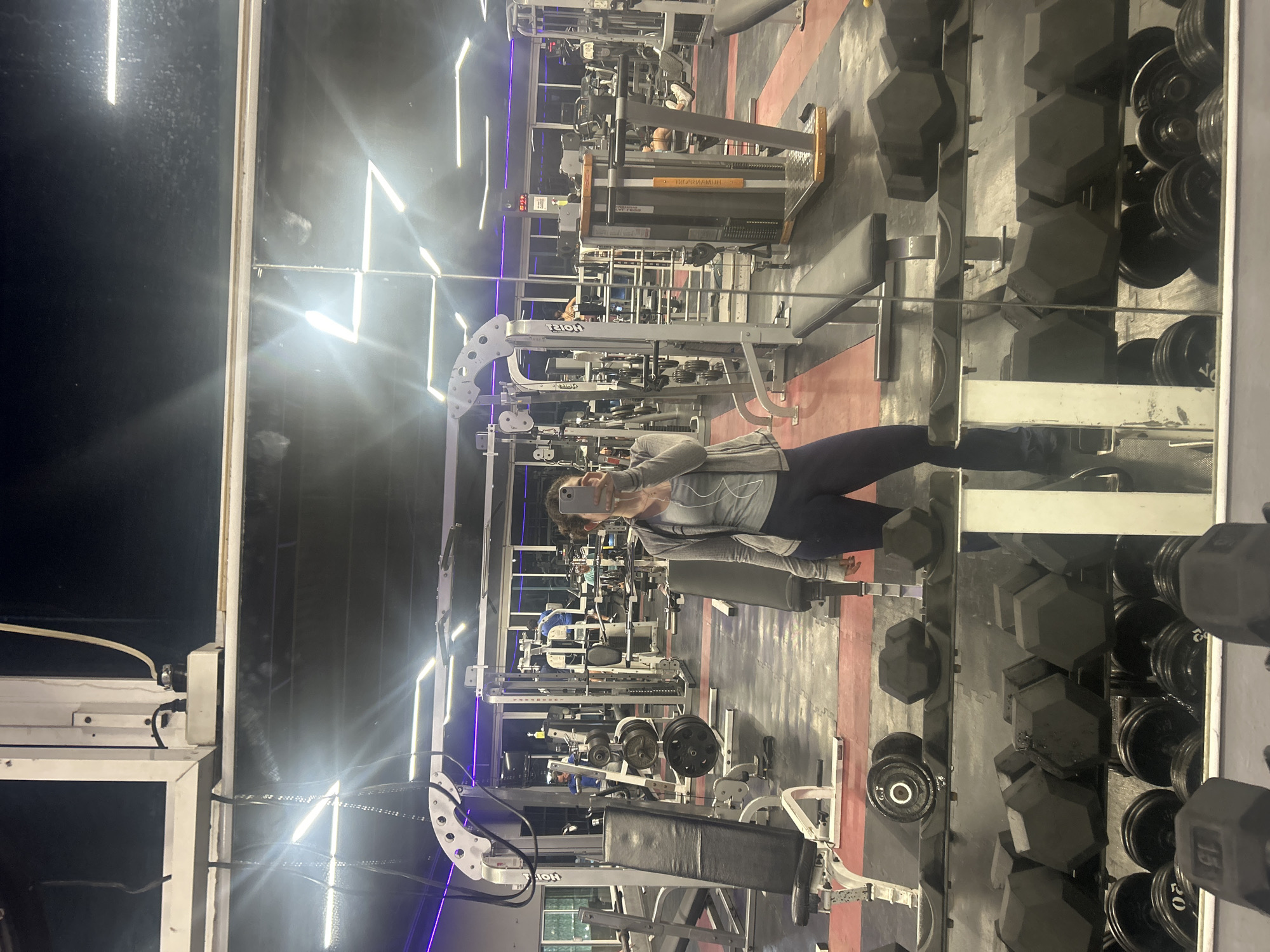 The image depicts a woman taking a selfie in a gym. The gym has various exercise machines, including weightlifting benches, pulley systems, and dumbbell racks. The floor has a red and grey color scheme. The location is in the city of Unknown, Unknown.