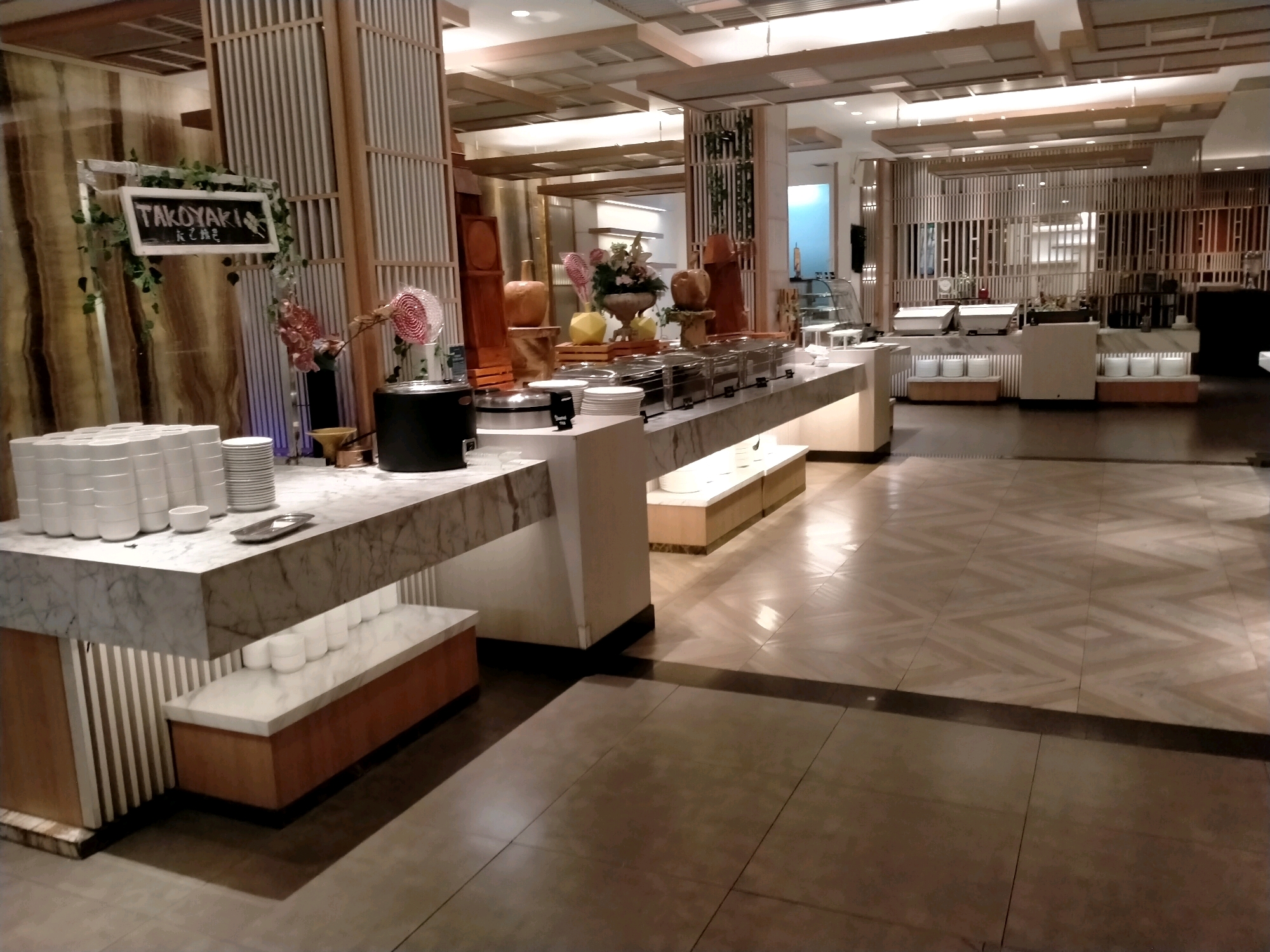 The image depicts an indoor buffet setting, likely in a hotel or restaurant, with a focus on food service areas. In the foreground, a marble-topped counter displays stacks of white plates and bowls, along with a small silver tray. To the left, more stacks of white plates are visible. A black appliance, possibly a warmer or cooker, is positioned behind the plates. Above the counter, a sign reads "TAKOKYAKI" in white lettering on a black background, adorned with green ivy. Further back, a buffet line features stainless steel food containers and decorative items such as a floral arrangement and carved wooden pieces. The flooring is a polished tile with a patterned design. The overall environment suggests a well-maintained dining area. There are no people visible in the image.