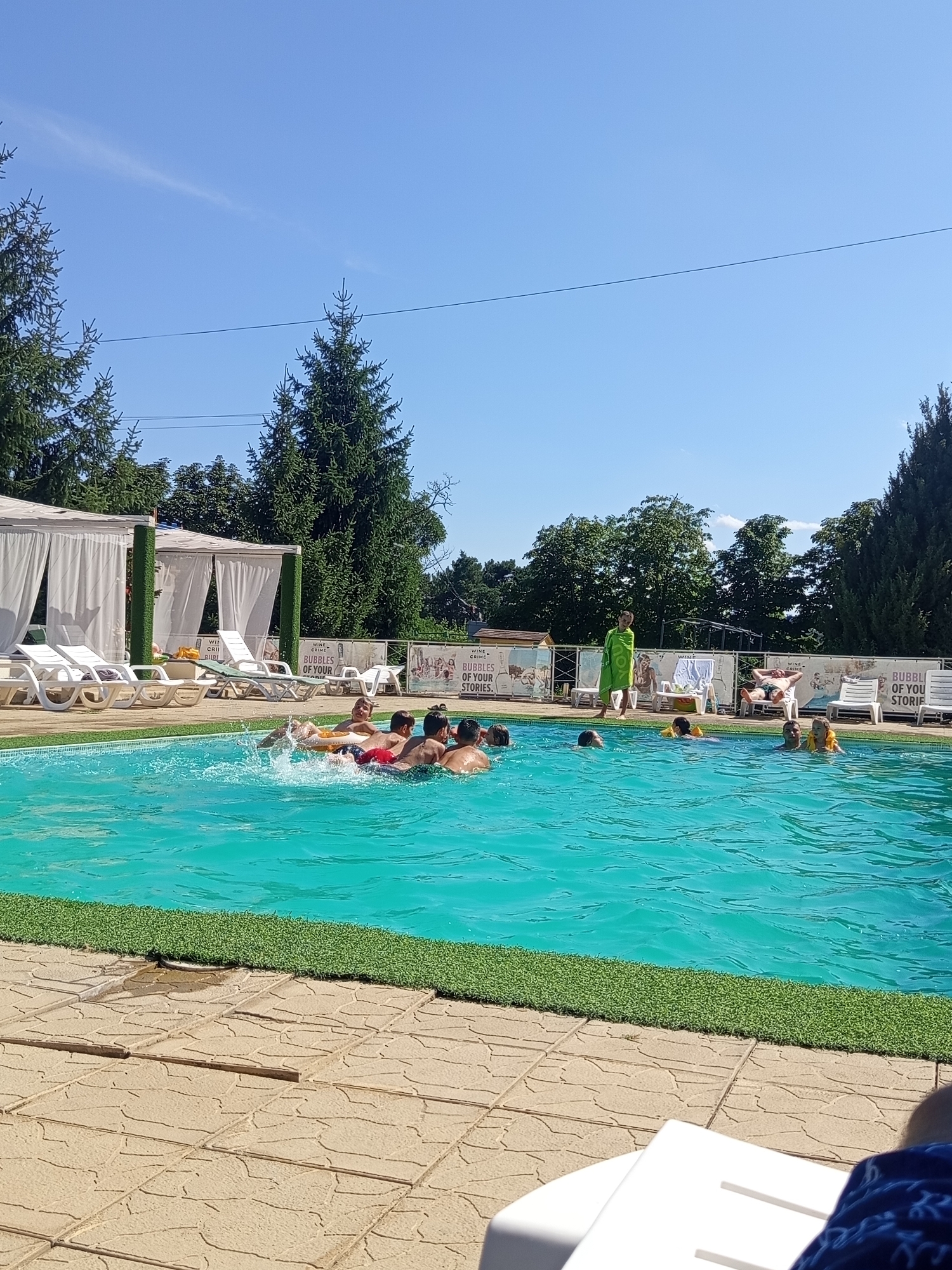 In Molovata Nouă, Moldova, a group of individuals are enjoying a sunny day at an outdoor swimming pool. The pool's water is a vibrant turquoise, with visible ripples and splashes indicating activity. Several people are in the water, with some clustered together and appearing to be interacting. One group of young individuals is on an inflatable, with splashes suggesting playful movement. Another person, wearing a green towel, stands by the pool. White lounge chairs are visible on the paved poolside, along with white cabanas with draped curtains. The area is bordered by artificial turf. In the background, mature green trees and a clear blue sky frame the scene. A banner with text is also present.