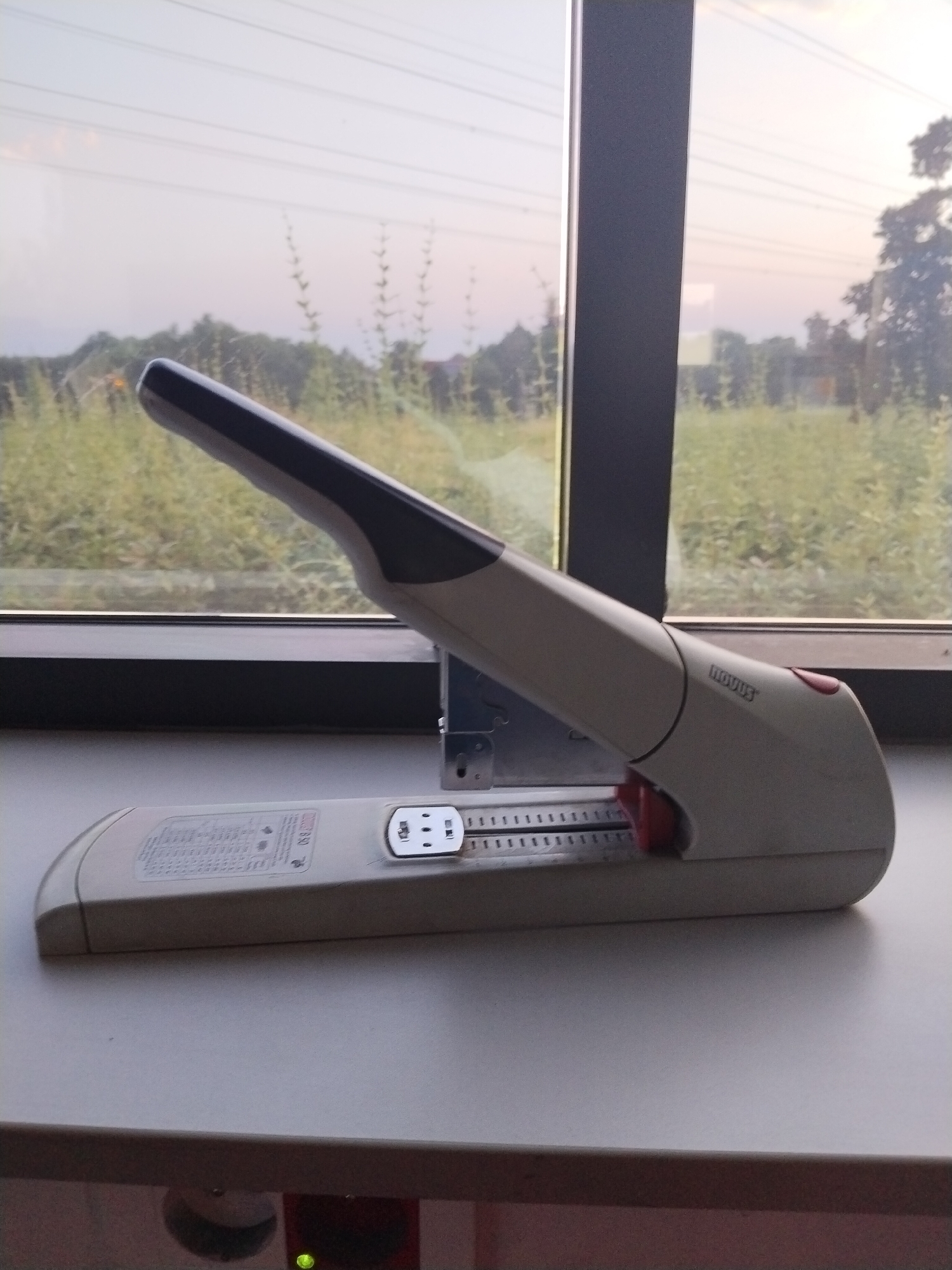 This image shows a stapler placed on a windowsill, with a blurred outdoor scene visible through the window. The stapler is a light beige color with a dark gray handle. A red lever is visible on the side of the stapler. Text on the stapler reads "NOVUS". The outdoor scene behind the window consists of green foliage and trees, suggesting a natural or rural setting. The sky is a pale, hazy blue, indicating it might be late afternoon or early morning. The lighting suggests a soft, diffused natural light. The overall impression is one of a quiet, everyday object set against a natural backdrop. There is no activity or event taking place; the image is a still-life composition. The location is identified as Nový Jičín, Czechia, though this context is not visually apparent within the image itself.