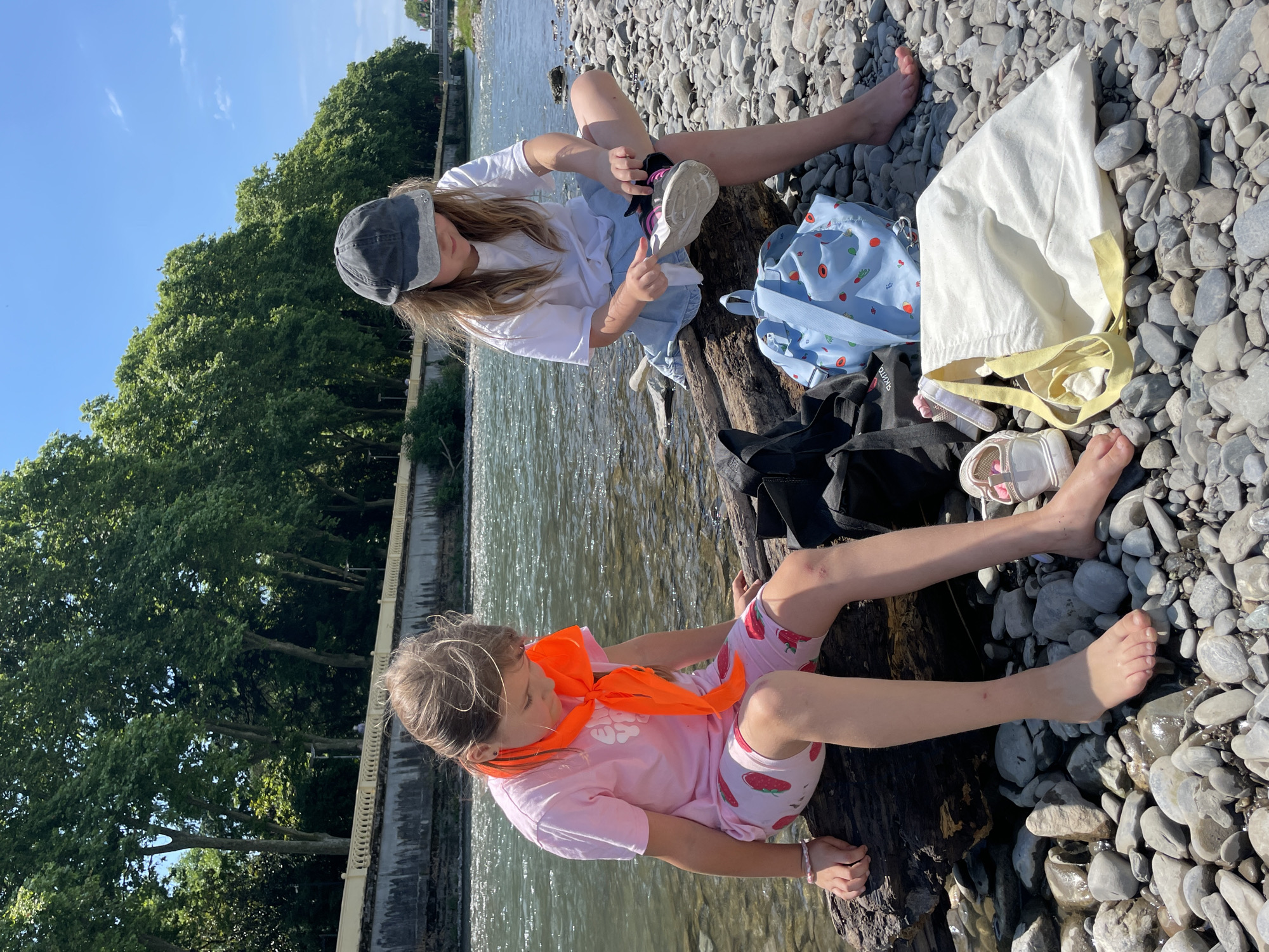 The image depicts two young girls sitting on a rocky shore next to a body of water, likely a river or lake. The sun is shining brightly, suggesting it's daytime, and the weather appears to be warm and pleasant.
The girl on the left is wearing a white t-shirt, blue denim shorts, and a dark grey baseball cap. She is actively engaged in putting on a white sneaker with pink laces. Beside her is a light blue backpack adorned with a strawberry pattern.
The girl on the right is wearing a pink t-shirt with a strawberry pattern and matching strawberry-patterned shorts. She has an orange bandana or scarf tied around her neck. She is sitting with her legs extended, bare feet resting on the rocky ground.
The ground is covered in smooth, grey pebbles. A large piece of driftwood serves as a natural bench for the girls. In the background, lush green trees line the opposite bank of the water, and a low concrete wall with a decorative railing is visible along the water's edge. A white tote bag with yellow straps is also present near the girl on the left.
The overall mood seems relaxed and summery, with the girls enjoying a day outdoors near the water. No visible text is discernible in the image. The location is identified as Sochi, Russia.