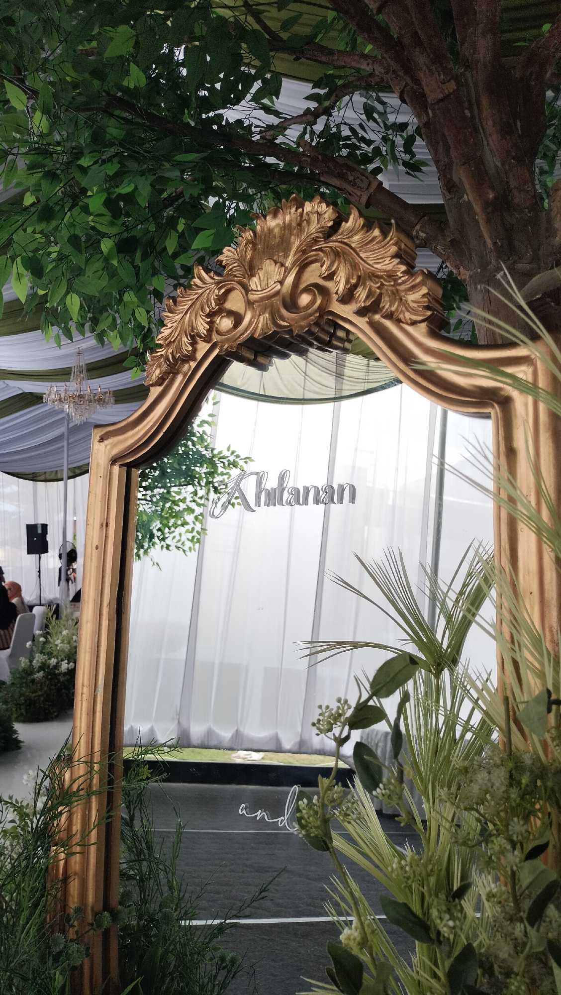 The image depicts a decorative mirror with a golden, ornate frame positioned outdoors, possibly at an event. The mirror reflects a backdrop of white drapes and greenery, suggesting an outdoor or semi-outdoor setup with artificial foliage and a possible tree trunk visible on the right. The reflection also shows the word "Ahlananan" in silver lettering on the mirror's surface, and further down, the word "and" in a similar style. In the background, white tent structures and subtle lighting are discernible. The foreground is dominated by lush green artificial plants, partially obscuring the lower part of the mirror and the ground. There are no discernible individuals or specific landmarks that would place this image in Lhokseumawe, Indonesia.