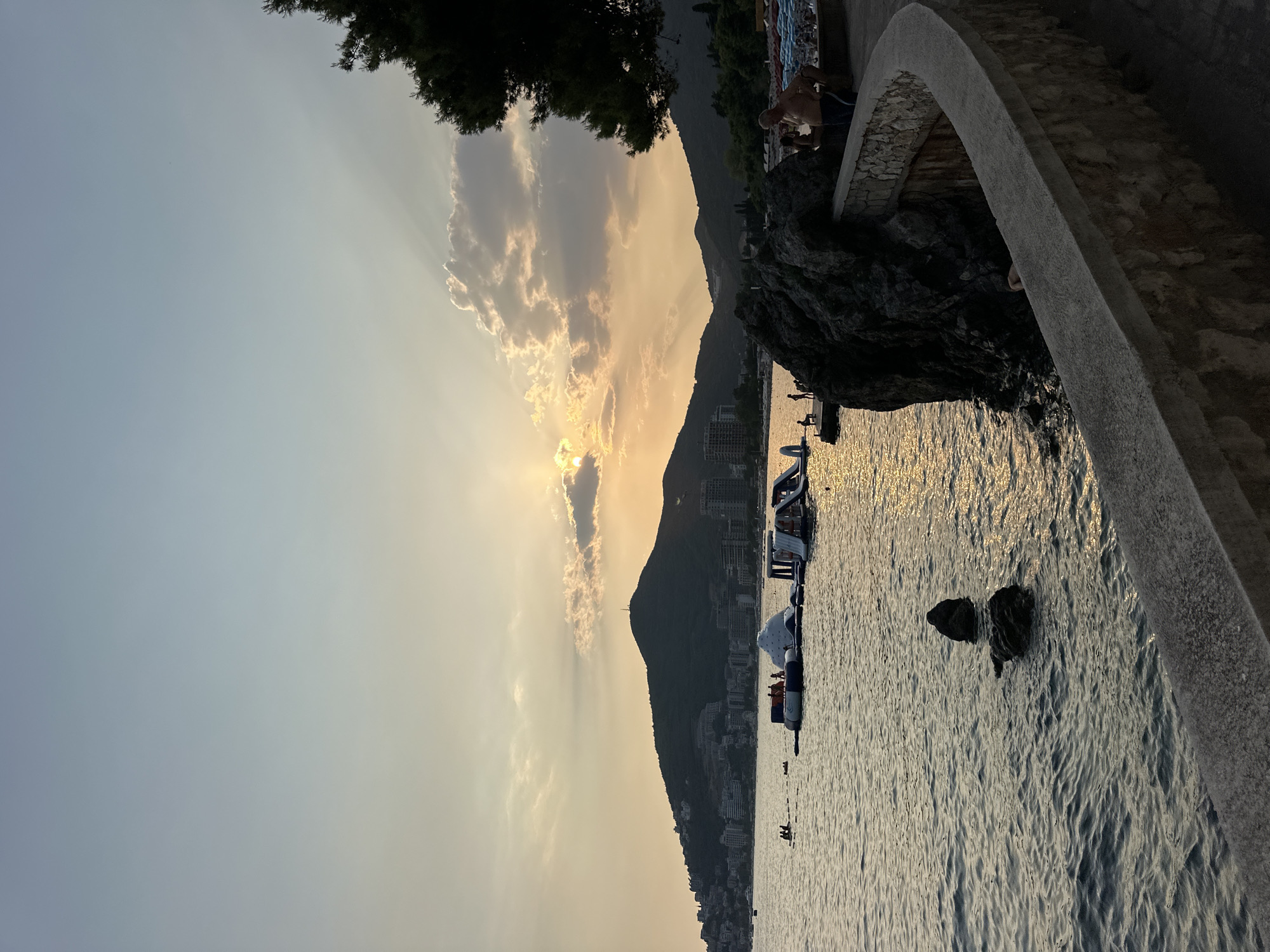 The image captures a serene sunset over the Adriatic Sea in Budva, Montenegro. The sun is partially obscured by clouds, casting a warm, golden glow across the sky and water. In the foreground, a curved stone wall separates a paved walkway from the sea. To the left, rocky outcrops break the surface of the water.
Further out in the bay, a floating water park with inflatable slides and structures is visible, with several people enjoying the amenities. In the middle ground, the coastline of Budva stretches out, lined with buildings and a prominent, tree-covered hill with a structure at its summit. Silhouetted against the sunset, the hills of Montenegro form a majestic backdrop to the scene.
On the walkway to the right, several individuals are present, some appearing to be resting or observing the view. The overall atmosphere is one of tranquil leisure as the day draws to a close.