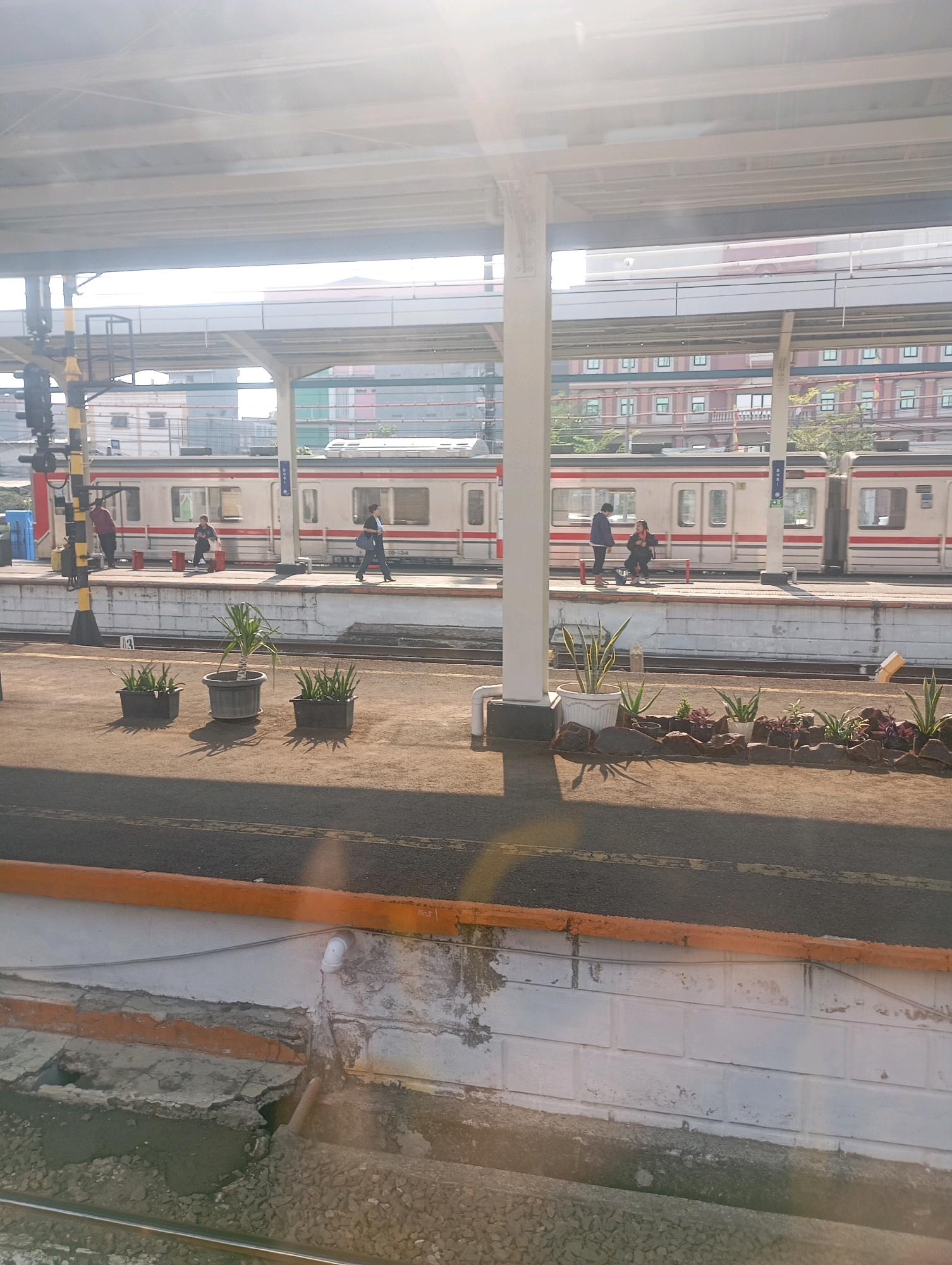 The image captures a sunlit train station platform in West Jakarta, Indonesia, likely during the morning. A passenger train, consisting of multiple cars with cream-colored bodies and red stripes, is stopped at the platform. Several individuals are visible on the platform: one person in a red shirt stands near the train, another in dark clothing is walking, and a third person in dark pants and a dark top is seated on a bench. The platform itself is constructed from concrete and lined with planters containing greenery, adding a touch of nature to the urban environment. In the background, multi-story buildings with visible windows suggest the presence of a city. The overhead canopy of the station provides shade, and the scene is illuminated by bright sunlight, creating strong shadows. The overall impression is a typical urban scene at a public transportation hub, bustling with quiet activity under a clear sky.
