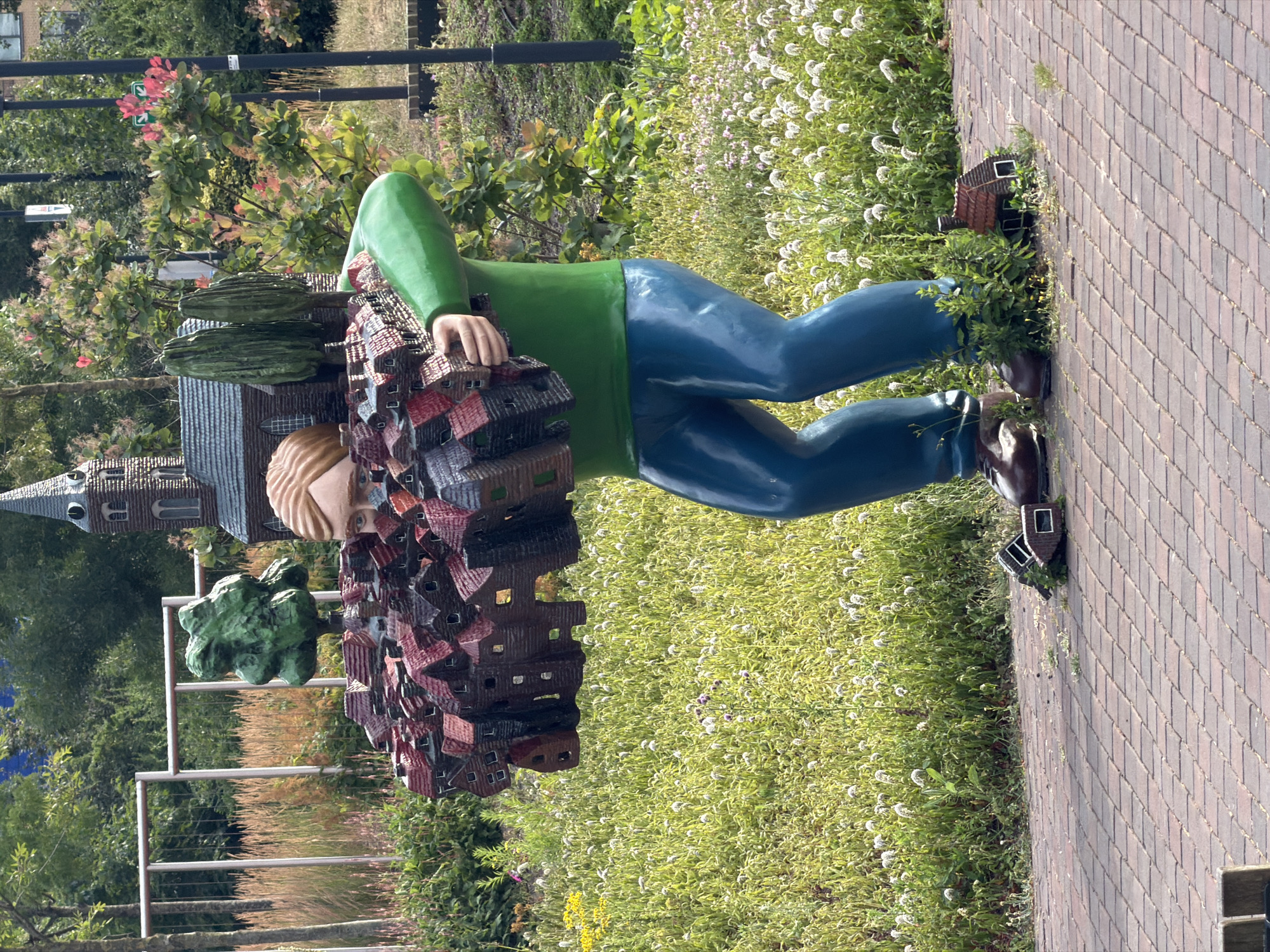 A full-body, low-angle shot captures a whimsical outdoor sculpture in Rijswijk, Netherlands. The sculpture depicts a stylized, cartoonish figure with smooth, rounded forms, wearing a bright green long-sleeved shirt and glossy blue pants. The figure is depicted in a crouching or bending posture, appearing to carry a large, dense cluster of miniature brick houses on its shoulders and arms. These miniature houses are multi-colored, primarily in shades of red and dark brown, with visible windows and roofs.
The figure's face is visible, with wide, alert eyes peeking out from behind the miniature cityscape. The figure's skin tone is a light brown. The sculpture is set against a backdrop of lush greenery, with a dense field of small white flowers and yellow wildflowers in the mid-ground. In the background, there are trees, a fence, and a building with a dark brick facade and a pointed steeple, resembling a church. The foreground consists of a brick-paved walkway. Two miniature houses, similar to those being carried by the sculpture, are placed on the ground near the sculpture's feet, one to the left and one to the right, partially obscured by vegetation. The overall scene suggests a playful and surreal artistic installation in a public outdoor space.