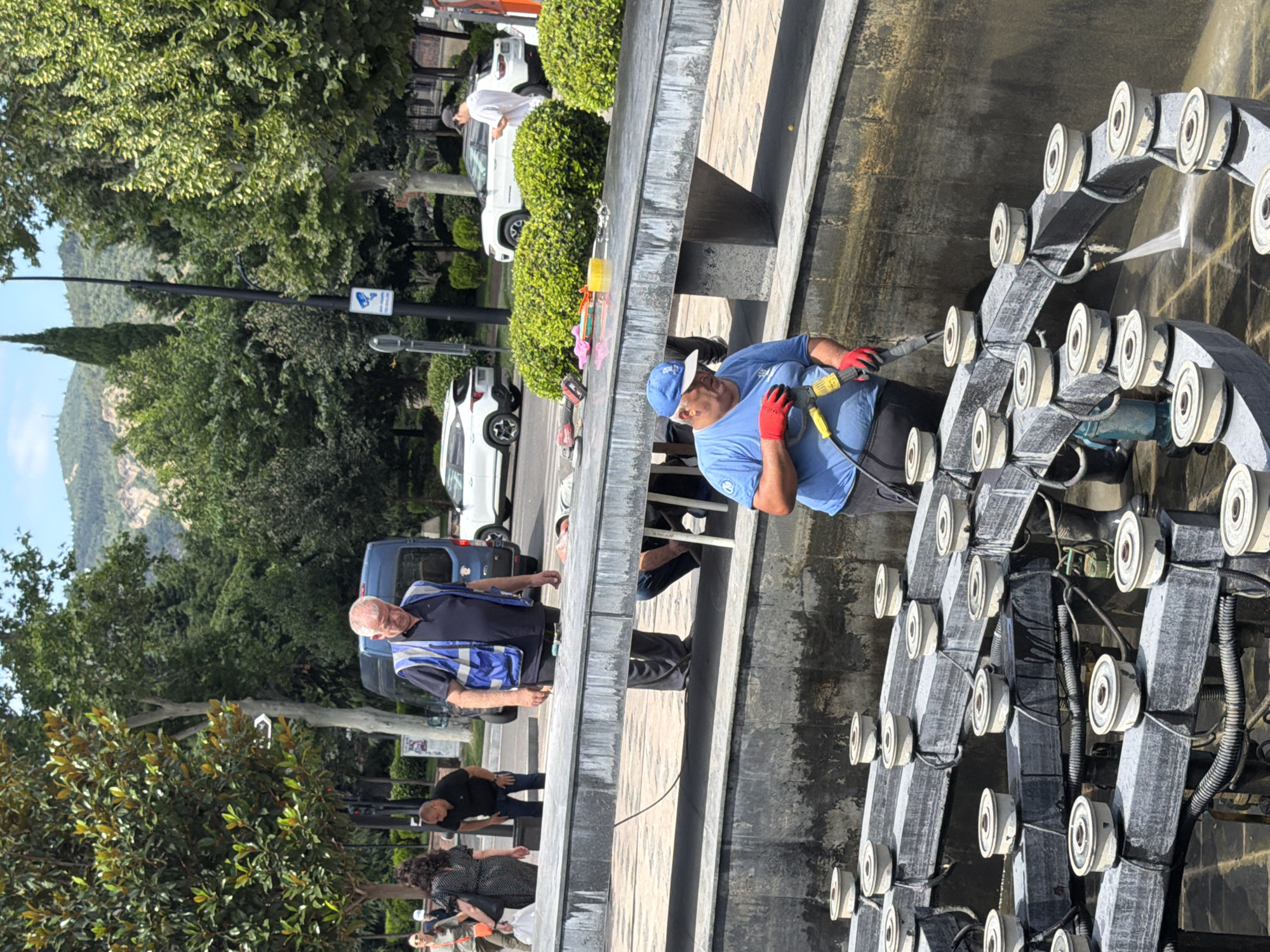The image captures a scene at the Dry Bridge Market in Tbilisi, Georgia, where workers are performing maintenance on a large, tiered fountain. In the foreground, a man wearing a blue t-shirt, a blue cap, and red gloves is operating a high-pressure washer, directing a stream of water onto the fountain's structure, which is filled with numerous circular nozzles and electrical wiring. Behind him, another worker in a blue vest and dark pants is standing near a blue utility van. Several other individuals are visible in the background, including a woman with dark curly hair carrying a plastic bag and a man in a black t-shirt. The surrounding environment includes trees, a street with cars, and distant hills characteristic of Tbilisi's landscape. The fountain appears to be made of concrete or stone, with a weathered, multi-layered design. The day is bright and sunny.