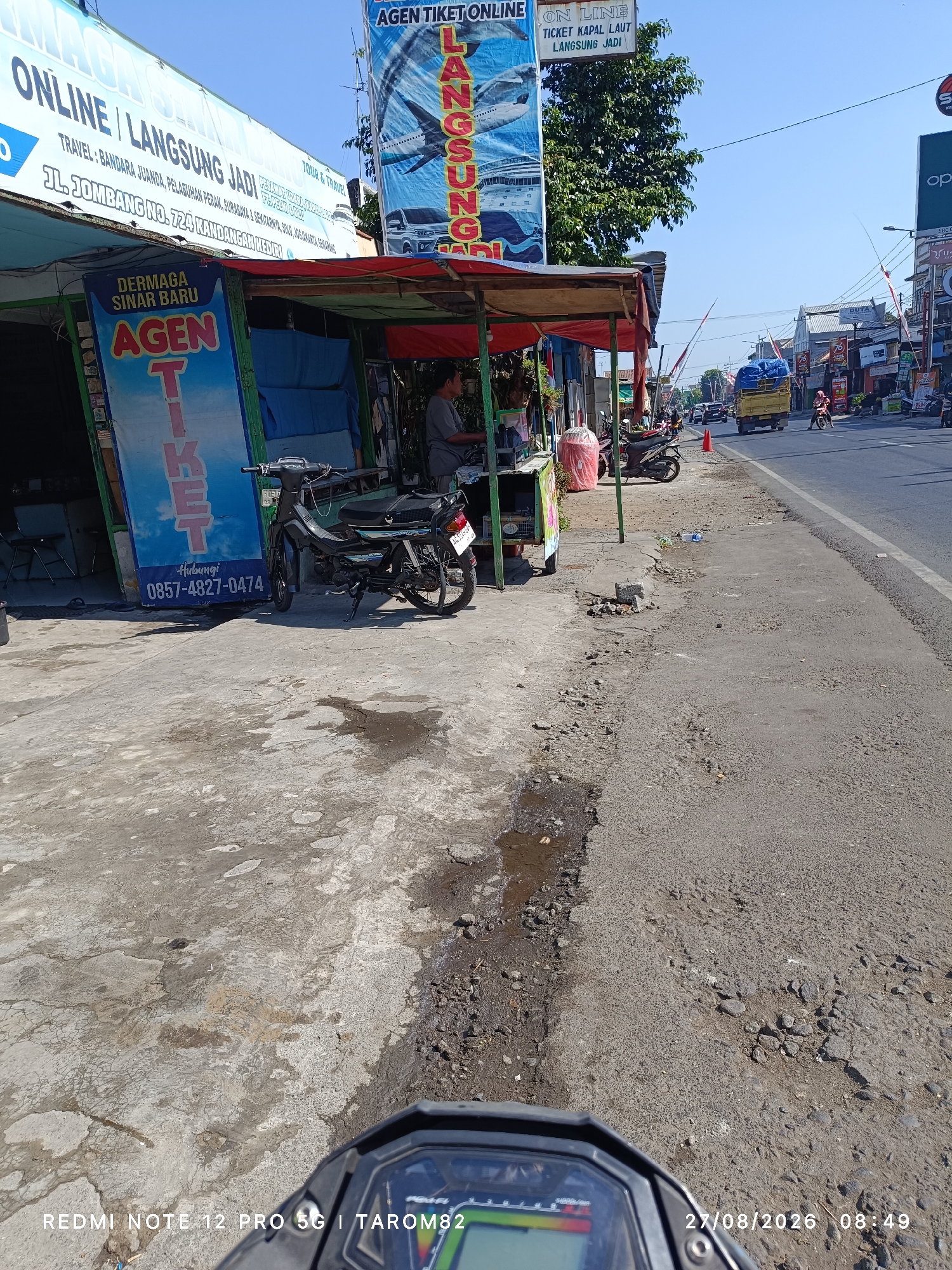 The image captures a street scene in Kandangan, Indonesia, featuring a small roadside stall. The stall appears to be a ticket agent for travel, with signs in Indonesian advertising "AGEN TIKET ON LINE" and "LANGSUNG JADI." One large banner is predominantly blue and white, displaying images of a cruise ship and airplanes, with text that reads "ONLINE / LANGSUNG JADI TRAVEL." Below this, another sign indicates "JL. JOMBANG NO. 724 KANDANGAN KEDIRI."
To the left of the main stall, a smaller blue and yellow sign is visible, stating "DERMAGA SINAR BARU AGEN TIKET" and providing a contact number: "0857-4827-0474." A motorcycle is parked in front of the stall. In the background, the street is lined with other shops and signs, and traffic, including a yellow truck and other vehicles, is visible. The road surface appears somewhat worn and uneven. The sky is clear and blue, suggesting a sunny day. A person is seen standing behind the stall. The bottom of the image shows a motorcycle's instrument panel and a timestamp indicating "27/08/2026 08:49" and "REDMI NOTE 12 PRO 5G | TAROM82."