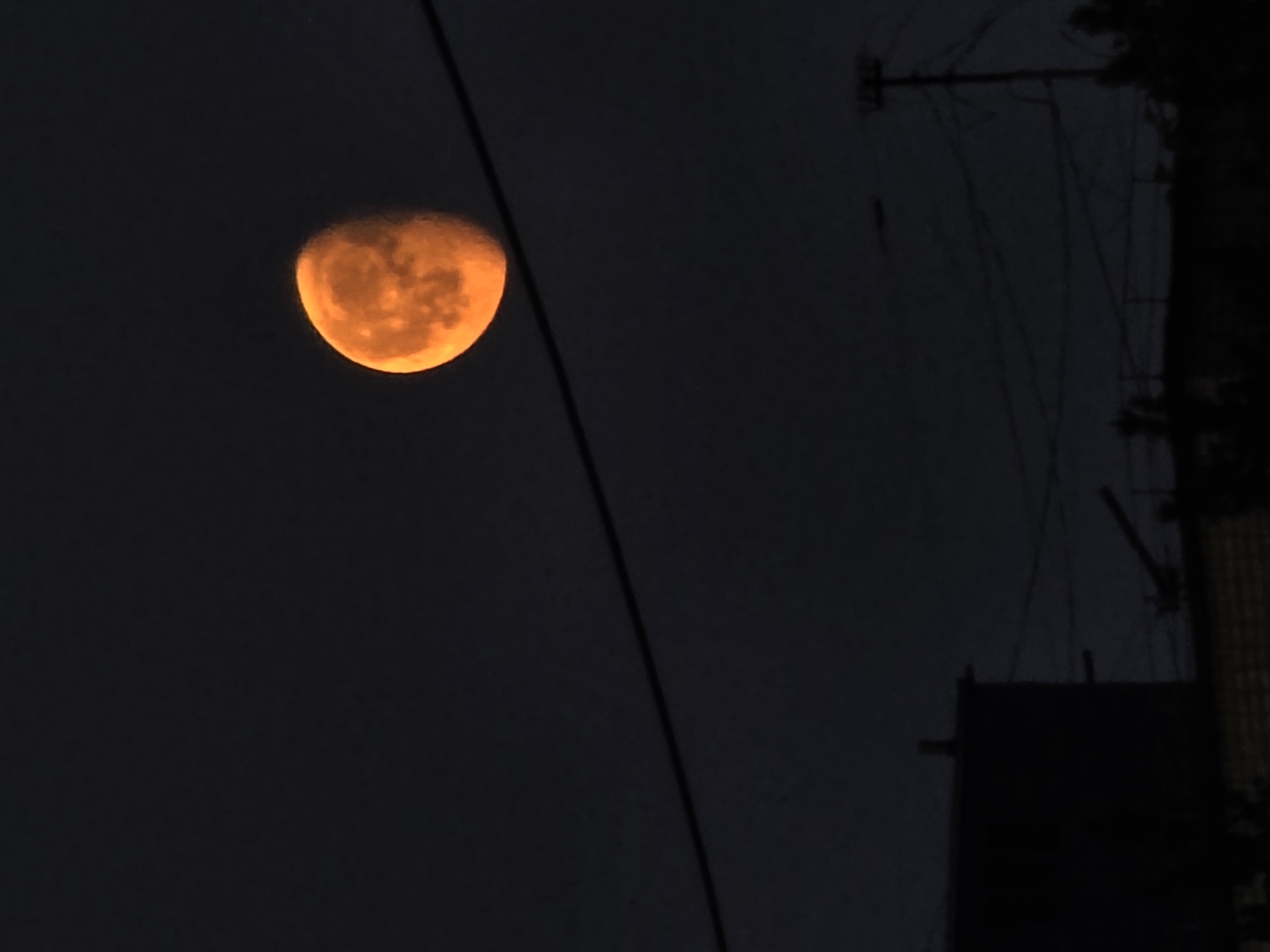 A dark, low-angle shot shows an orange moon in the night sky. The moon is not fully illuminated, appearing as a half-crescent shape with detailed surface textures. A power line diagonally crosses the frame from upper left to mid-right. In the darker right side of the image, there's a silhouette of what seems to be a utility pole or structure with multiple wires extending from it. A building or structure is partially visible on the right edge. This image was taken in Chaikovsky, Russia.