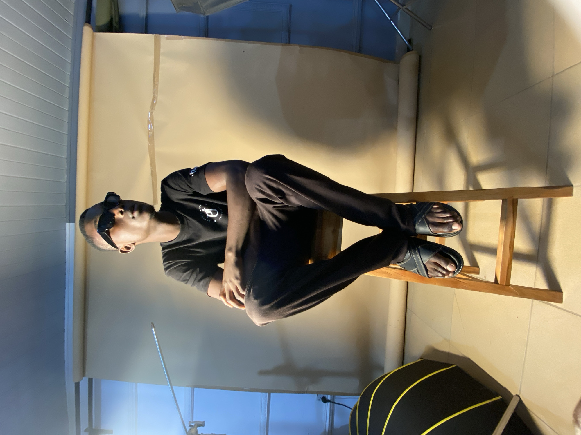 A person is sitting on a wooden chair. They are wearing a black t-shirt and black pants, with black sandals on their feet. The person has on dark sunglasses. The background appears to be a studio setting with a light-colored backdrop. There are also some rolled-up backdrops and studio equipment visible. The lighting suggests it is indoors, possibly during the day or evening. There is a black object with yellow stripes in the foreground, which could be part of the studio setup. The overall mood is relaxed and casual. The visible text on the t-shirt is not legible. The location context is Bukuru, Nigeria.
