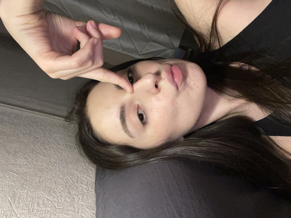 An indoor, close-angle selfie features a female subject with long, dark brown hair and fair skin. She is positioned slightly left of center, looking directly at the camera with her lips pursed into a "kiss" expression. Her right index finger presses against the bridge of her nose, while her other fingers are curled. She wears a dark-colored tank top. The background includes a dark grey pillow or cushion to her left, a light grey textured wall behind her, and a grey curtain with vertical folds on the right. No discernible landmarks or features in the image identify its specific location within Moscow, Russia, or any other city.