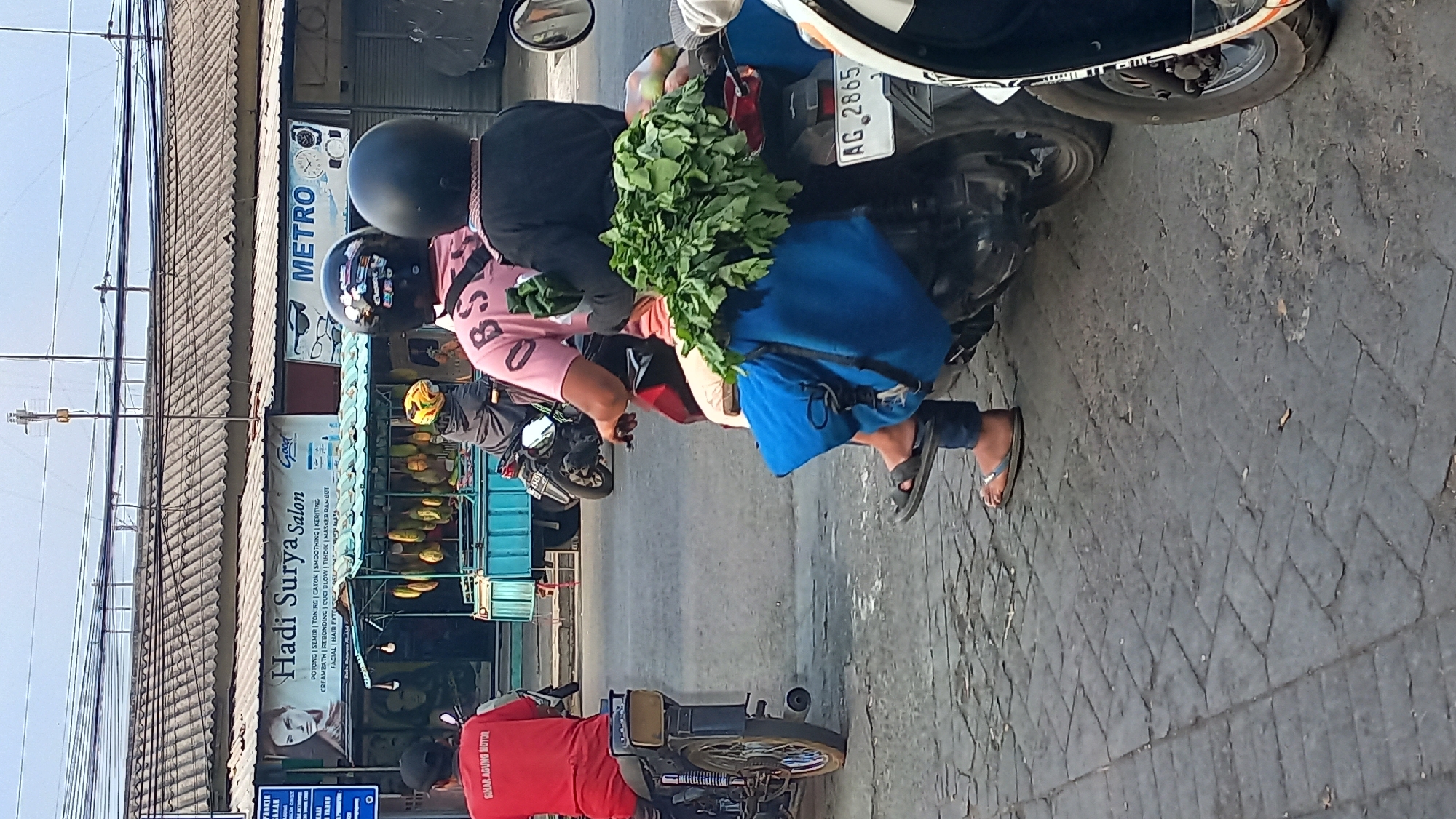 The image depicts a street scene in Mangunsari, Indonesia, where two motorcycles are parked next to each other on a paved road. On the left, a person wearing a red t-shirt with the text "SINAR AGUNG MOTOR" is seated on a motorcycle. In the center, another motorcycle carries two individuals. The person in front is wearing a pink t-shirt with the text "OBS" and a black helmet. Behind them, a person is wearing a black helmet and a black garment. A large bunch of green leafy vegetables is strapped to the back of this motorcycle, along with a blue bag. To the right, part of a third motorcycle is visible, with a white, black, and orange stripe on its side panel. In the background, a building with a sign that reads "Hadi Surya Salon" is visible, along with other businesses. A blue food cart with hanging produce, possibly durian, is parked in front of the salon. The sky is overcast, and power lines are strung across the top of the image.