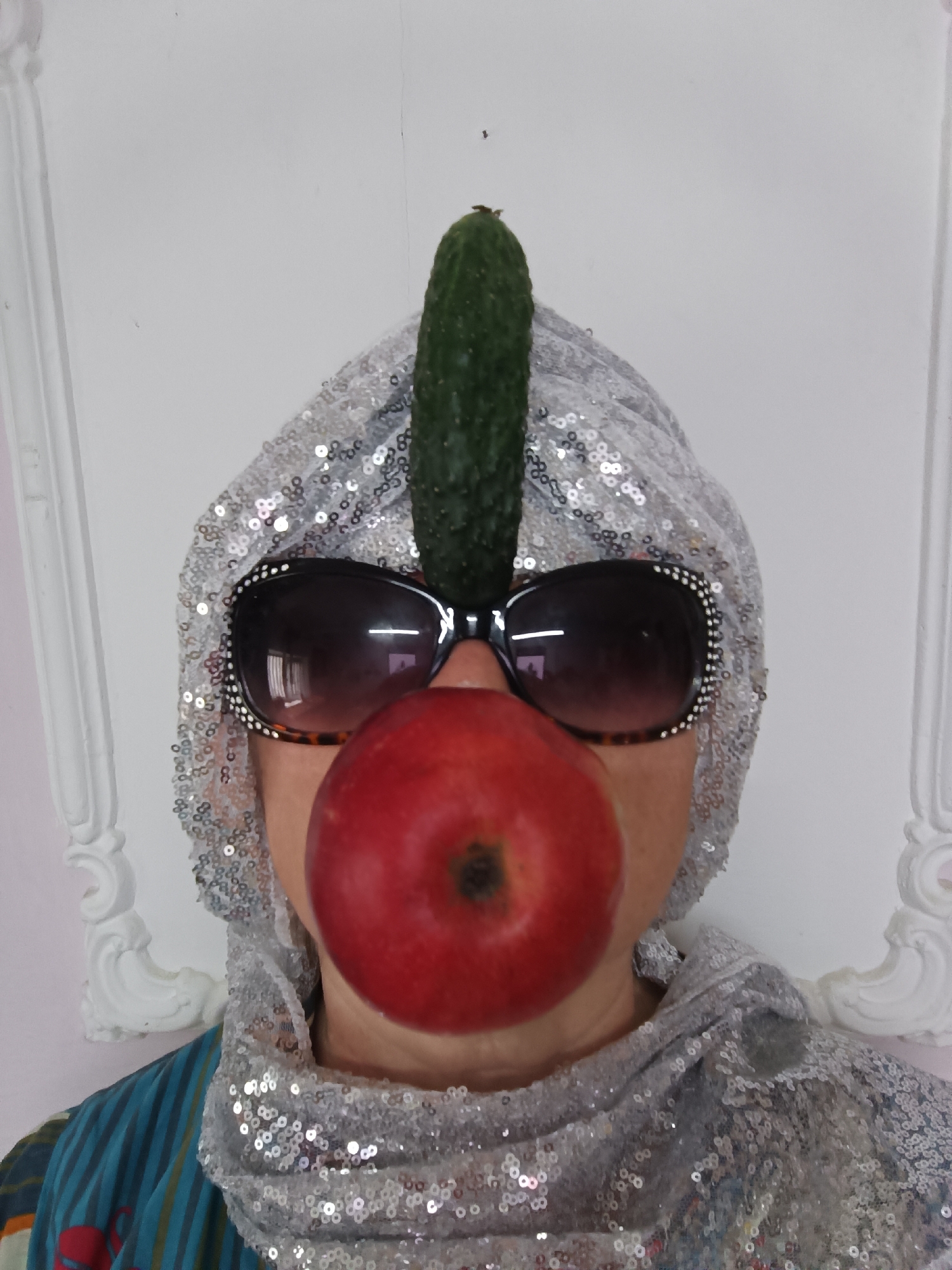 A person is standing in front of a white wall with decorative molding. They are wearing a sequined headscarf and a sequined scarf around their neck. Oversized sunglasses with rhinestones are perched on their nose, and a red apple is held in their mouth, obscuring it. A cucumber stands vertically on top of their head, balanced between the sunglasses and the headscarf. The person's clothing consists of a striped shirt with blue, green, and orange colors, and a red design visible on the sleeve. The image appears to have been taken indoors. There is no indication of the specific location within Tyshky, Ukraine.