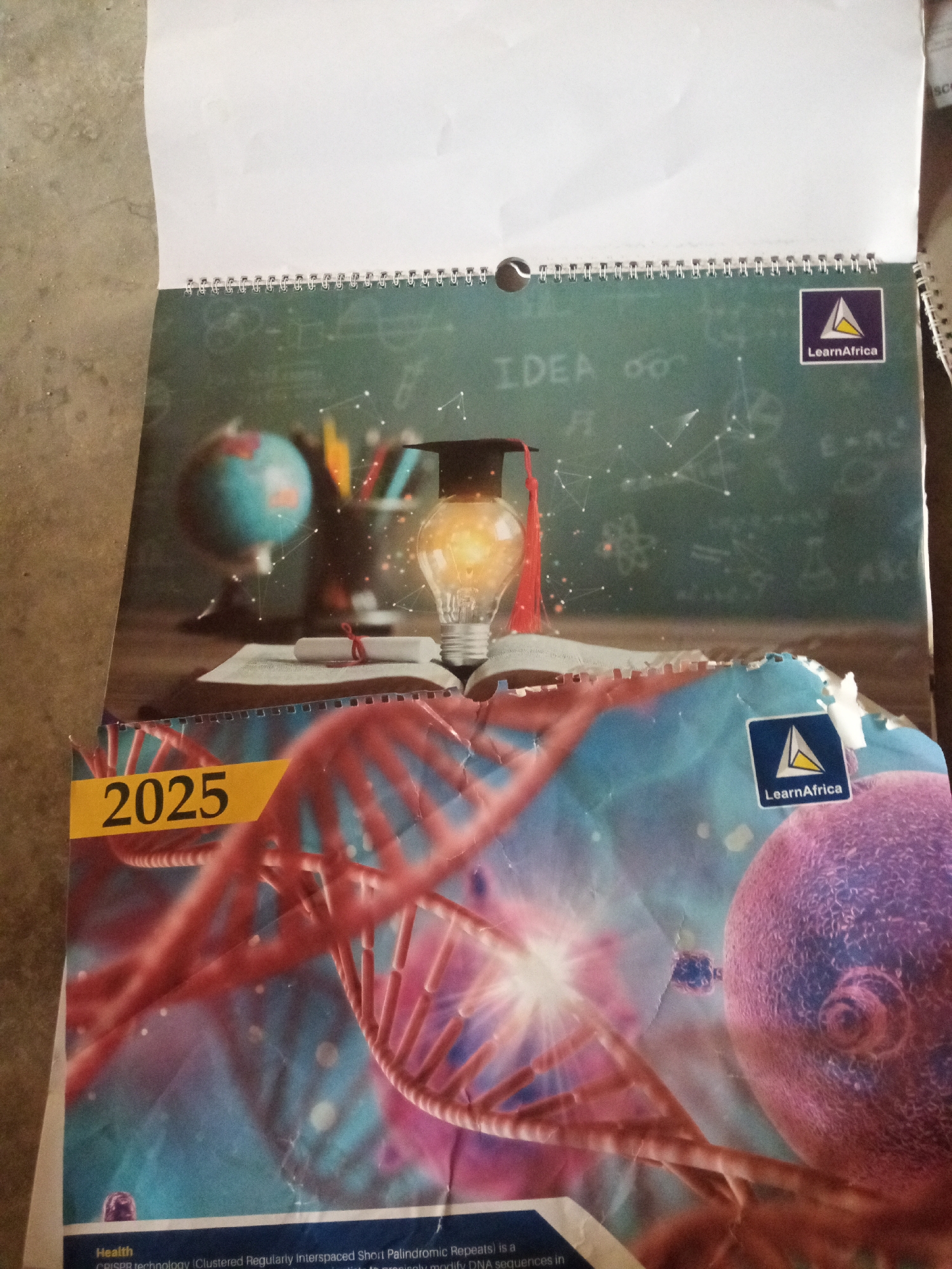 The image is a photograph of a spiral-bound notebook or calendar, possibly a school or educational planner, with a cover featuring scientific and educational imagery. The cover design includes a DNA strand, a depiction of a cell, a lightbulb symbolizing an idea, a globe, a graduation cap, and a book. The number "2025" is prominently displayed in yellow. There are two logos that appear to be for "LearnAfrica." The notebook is placed on a textured, possibly concrete surface. The lighting suggests it is indoors or in a shaded outdoor area. There is no indication of the city of Jalingo, Nigeria, or any people present in the image.