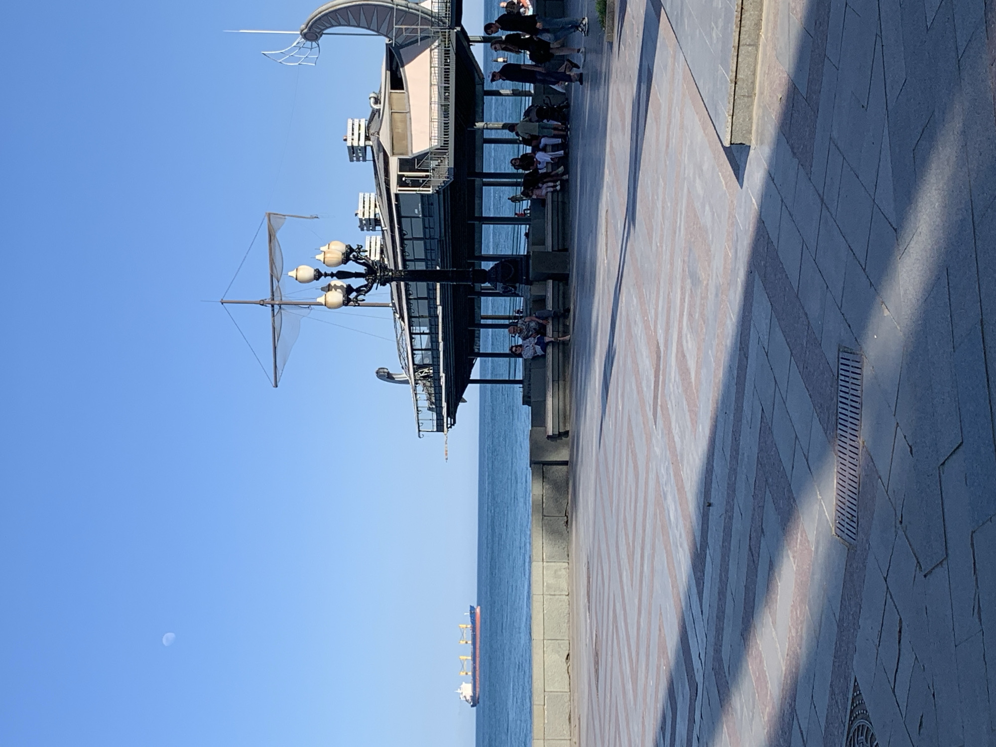 Here is a description of the image:
The image depicts a waterfront scene in an unidentified city. A pier structure extends over the sea, with people visible sitting and standing on it. The pier features a distinctive architectural design with multiple levels and a decorative canopy. A ship can be seen on the horizon. The sky is clear and blue, with a faint, pale crescent moon visible. The foreground shows a tiled walkway with a pattern of grey and beige. Shadows are cast across the walkway.